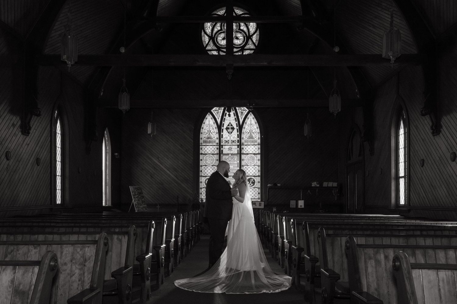 Black and white photo of a bride and groom standing at the altar of a historic wooden church with stained glass windows.