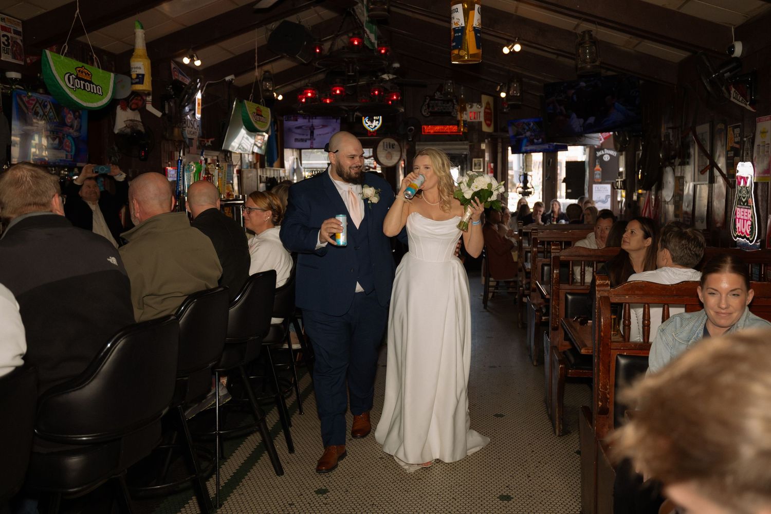 Bride and groom walking through a rustic bar venue during their wedding reception, surrounded by seated guests.