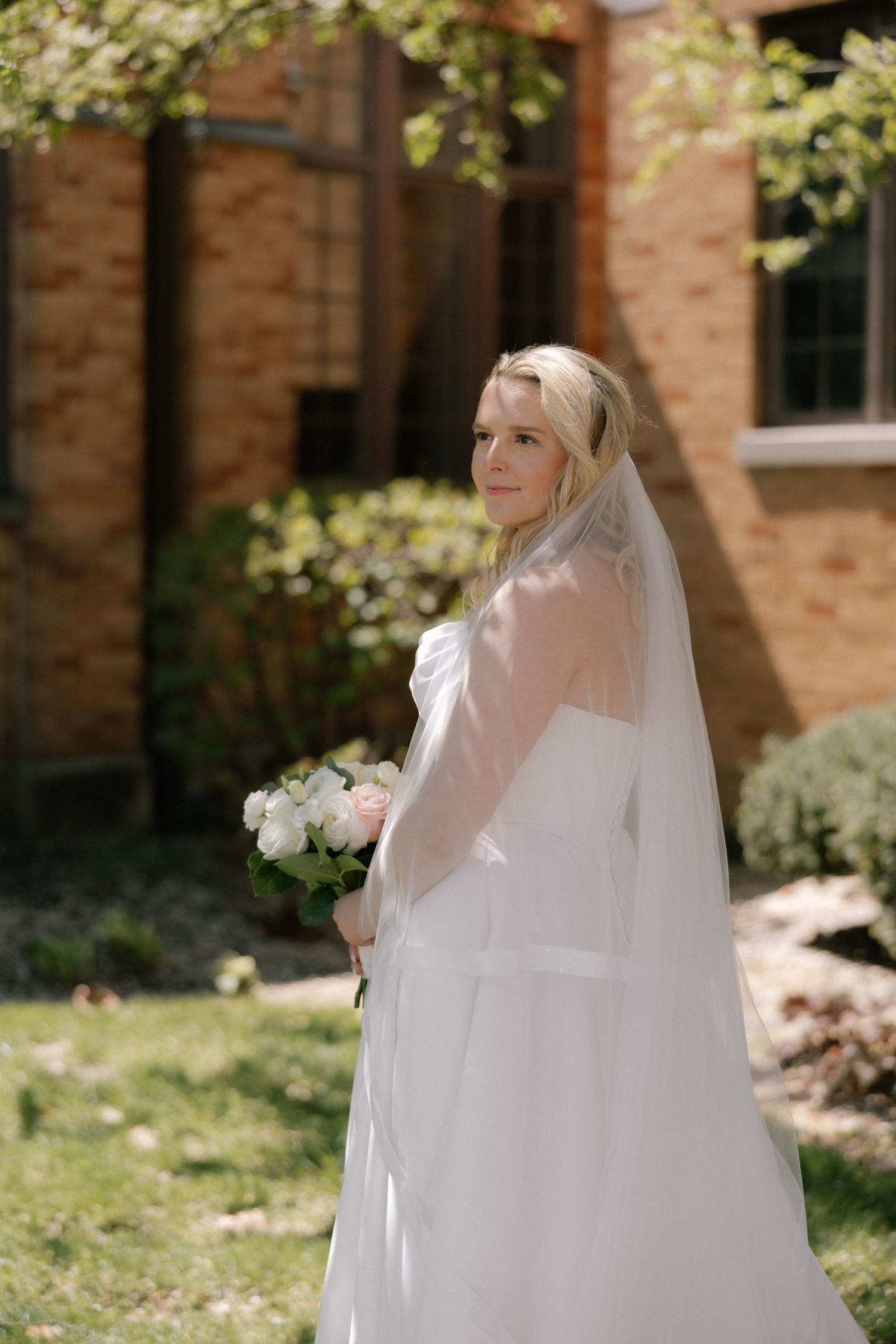 Bride in white gown and veil holds bouquet outdoors near brick building on sunny wedding day.