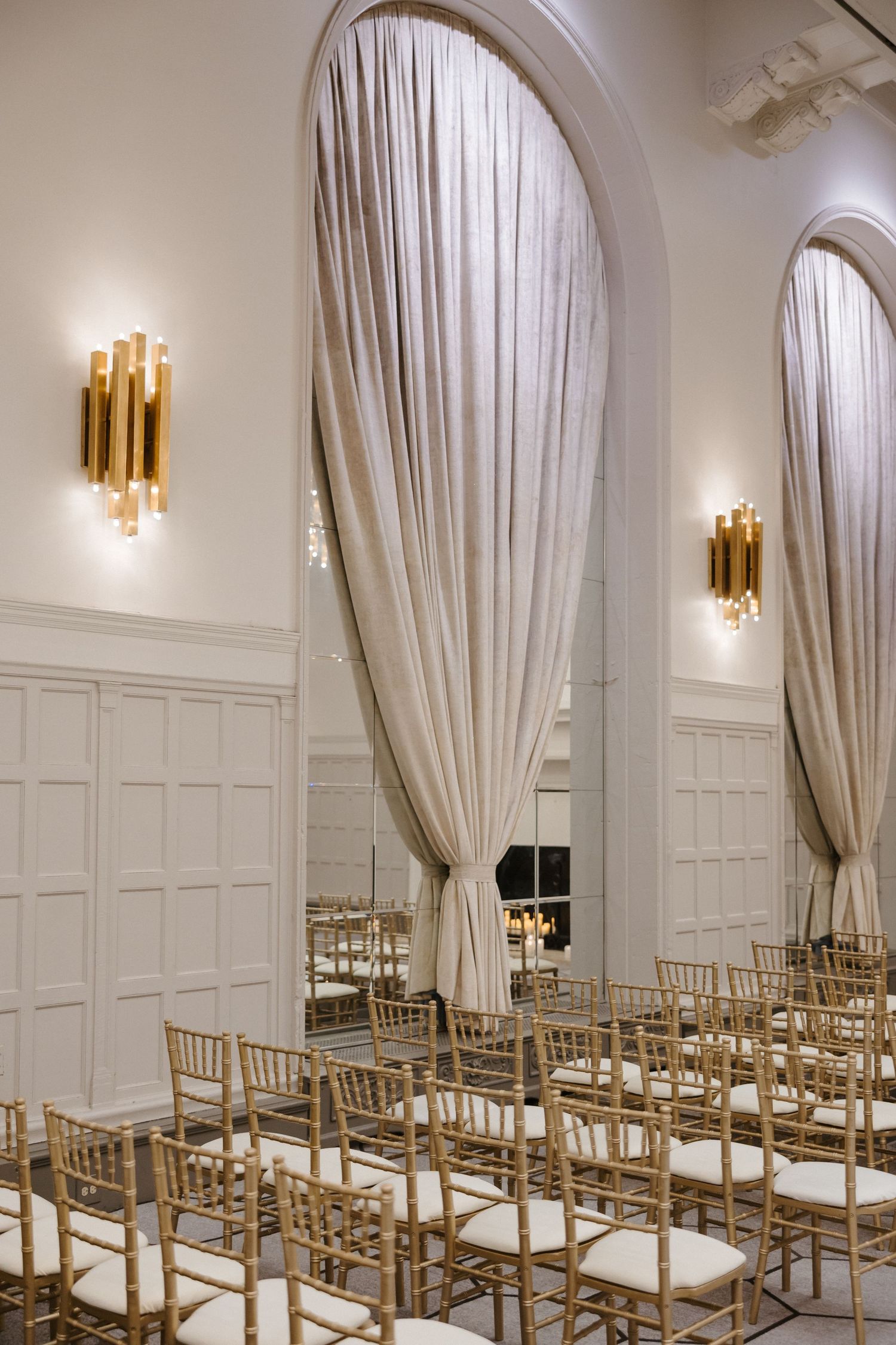 Elegant wedding ceremony venue with gold chiavari chairs, draped arched windows, and gold wall sconces on white paneled walls.
