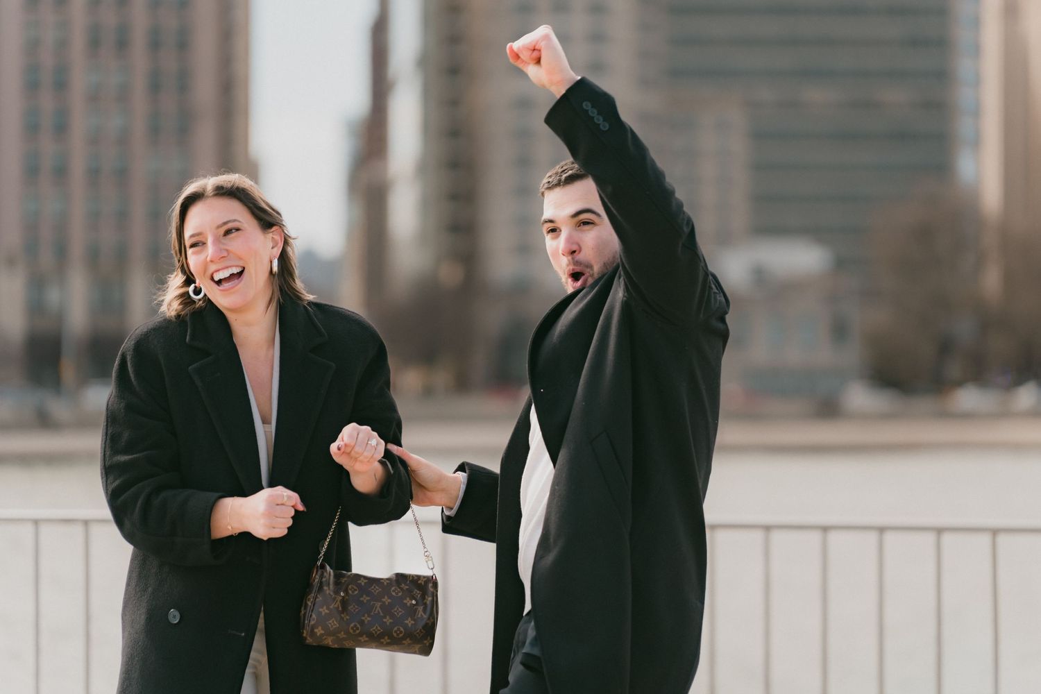 Joyful couple celebrating engagement outdoors in Chicago, woman laughing and man raising fist triumphantly.
