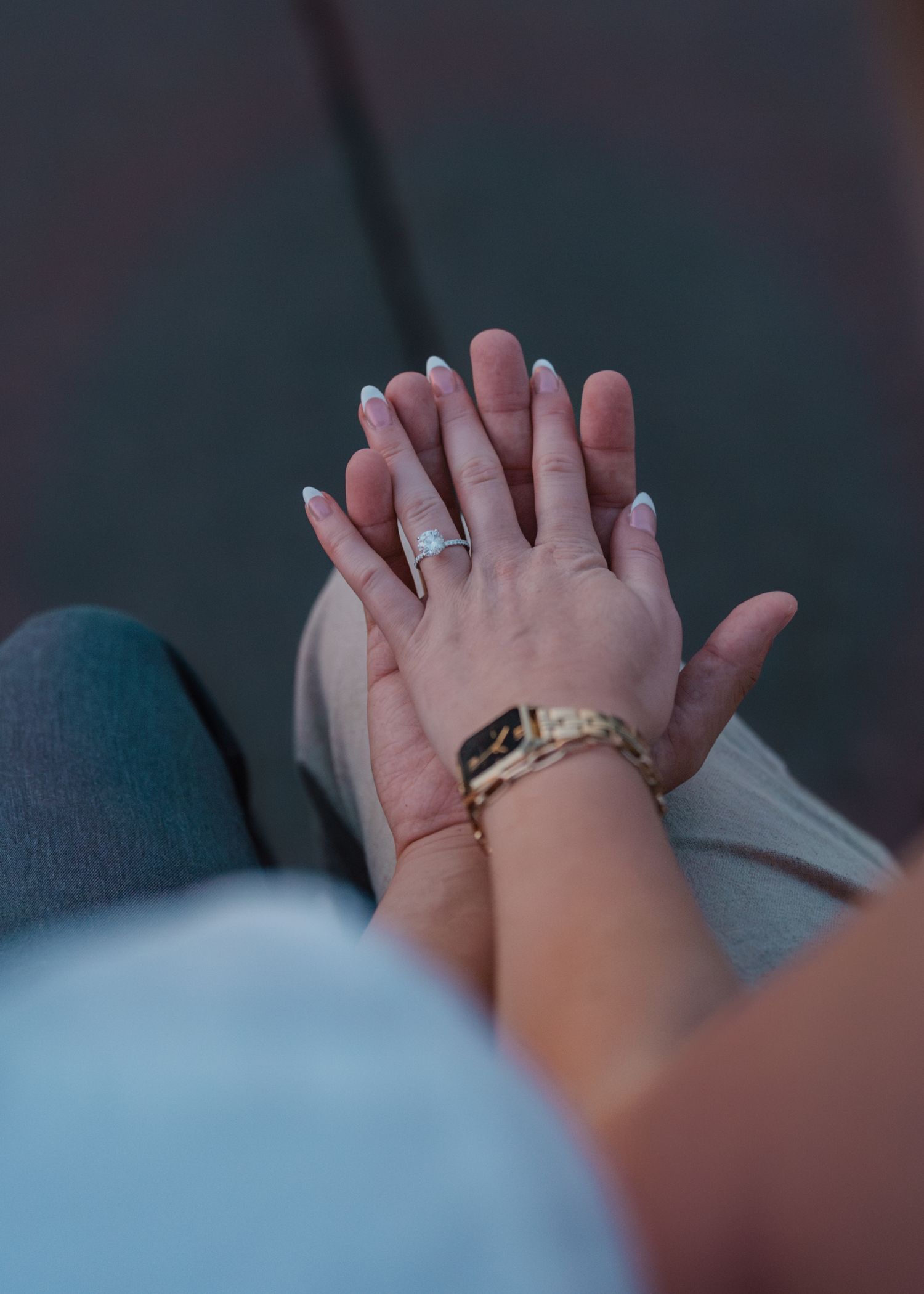 Two hands gently stacked together, one wearing a diamond engagement ring and a gold Apple Watch band.
