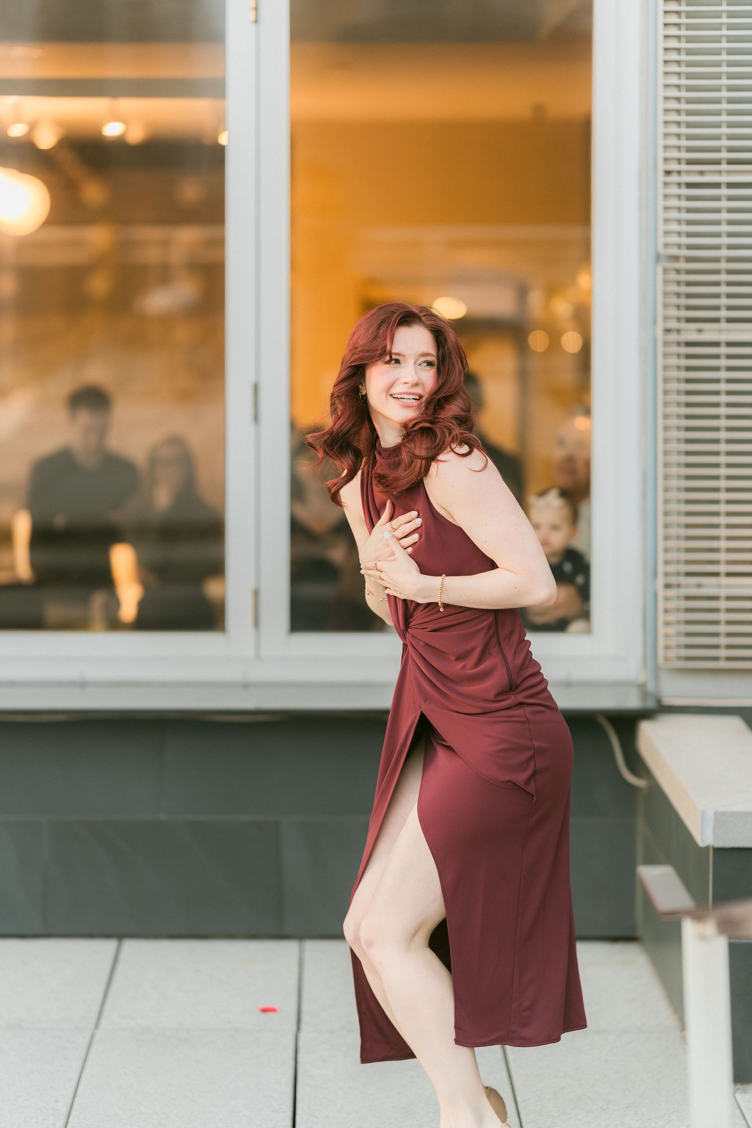 Elegant woman in a burgundy wrap dress posing outdoors near a glass building at golden hour.