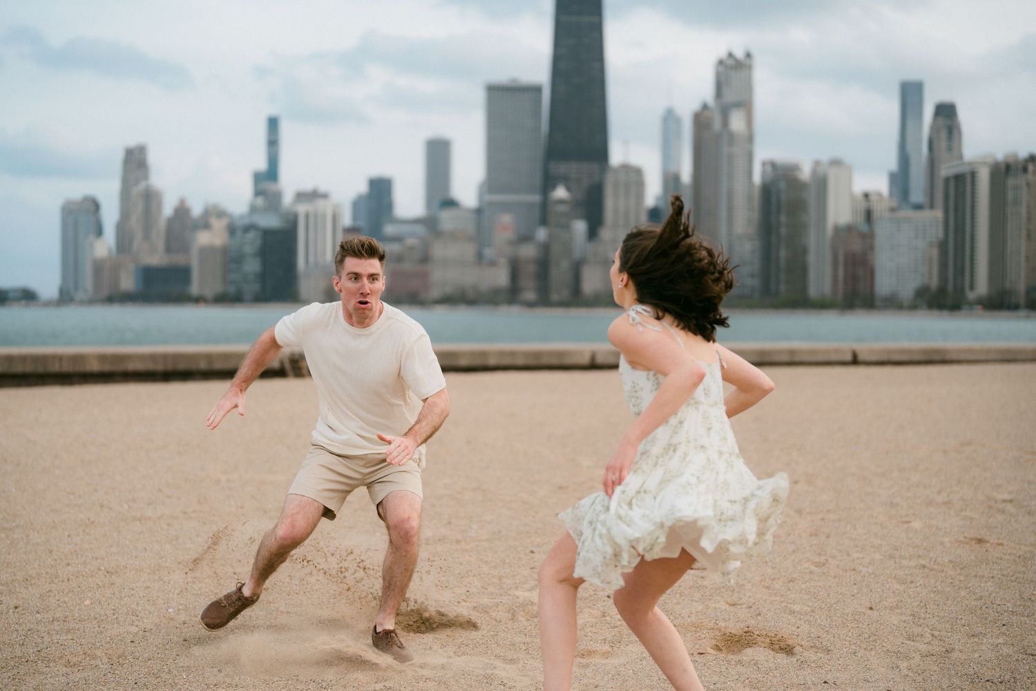 Couple playfully chasing each other on sandy beach with Chicago skyline behind them.