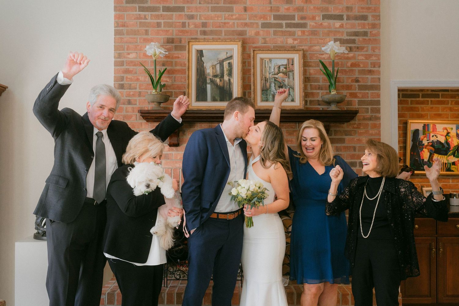Bride and groom kissing surrounded by cheering family members in front of brick fireplace.