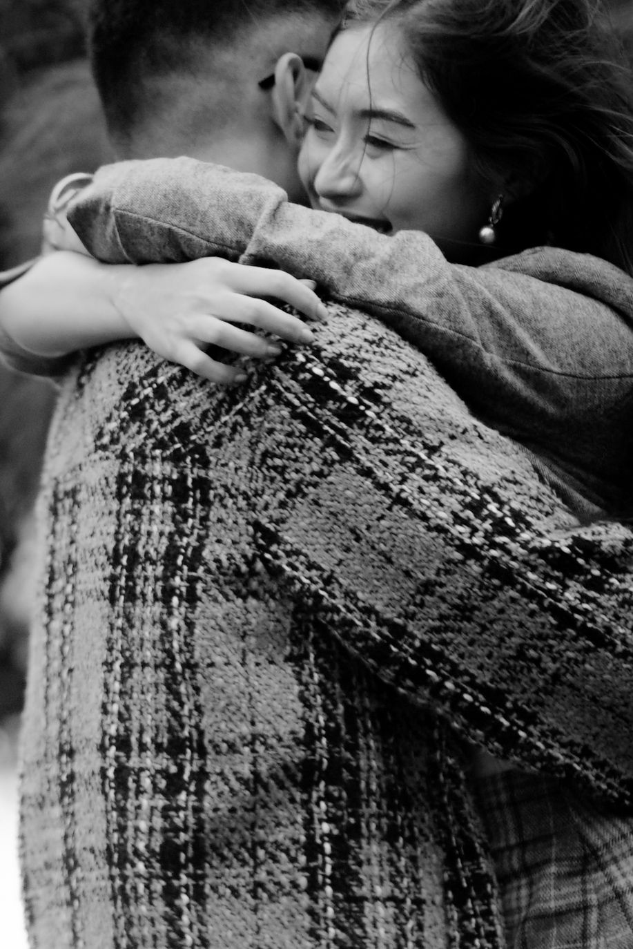 Black and white photo of a smiling woman hugging a man in a plaid coat, capturing a joyful embrace.