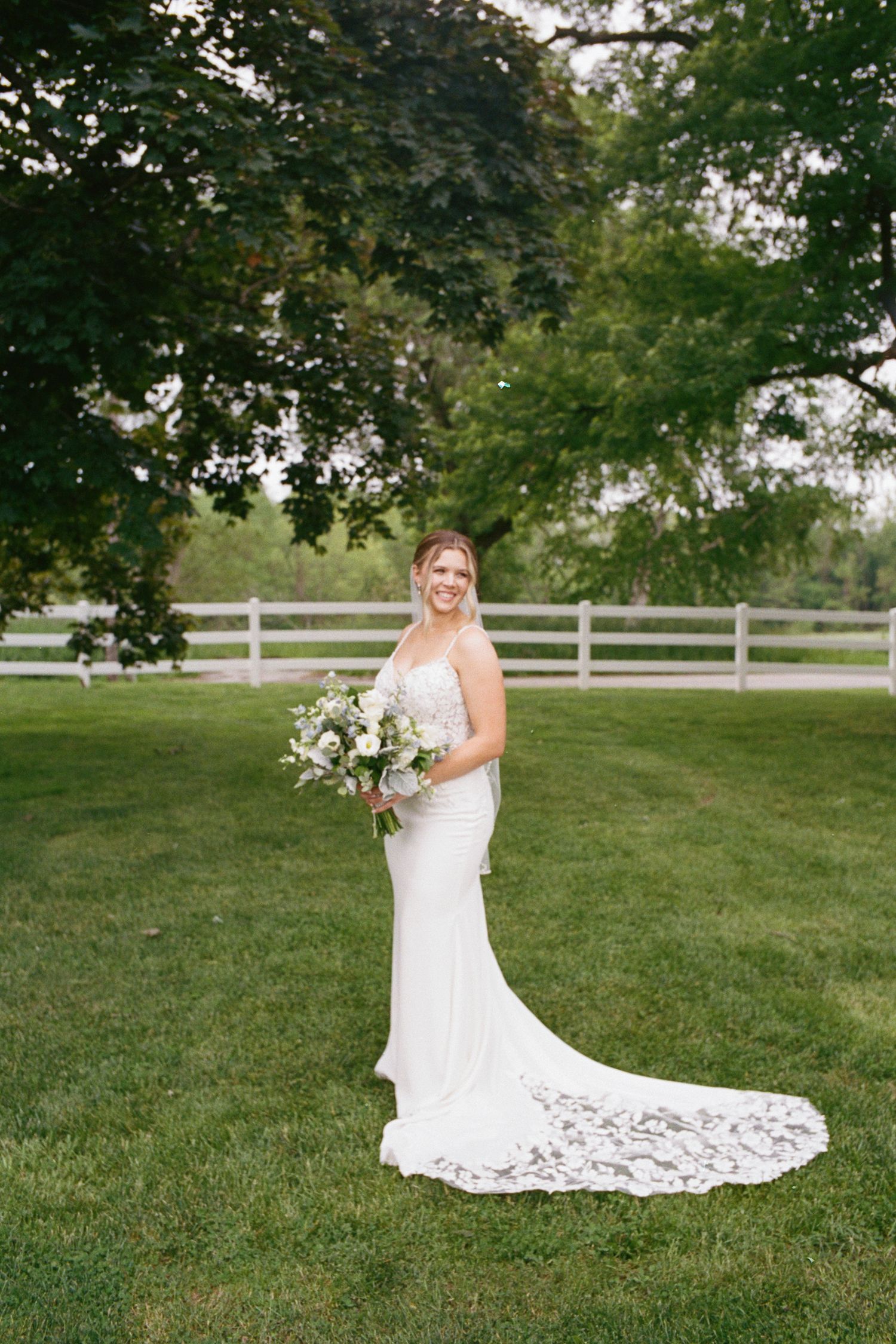 Bride in a white lace train gown holds a bouquet on green lawn with white fence and trees backdrop.