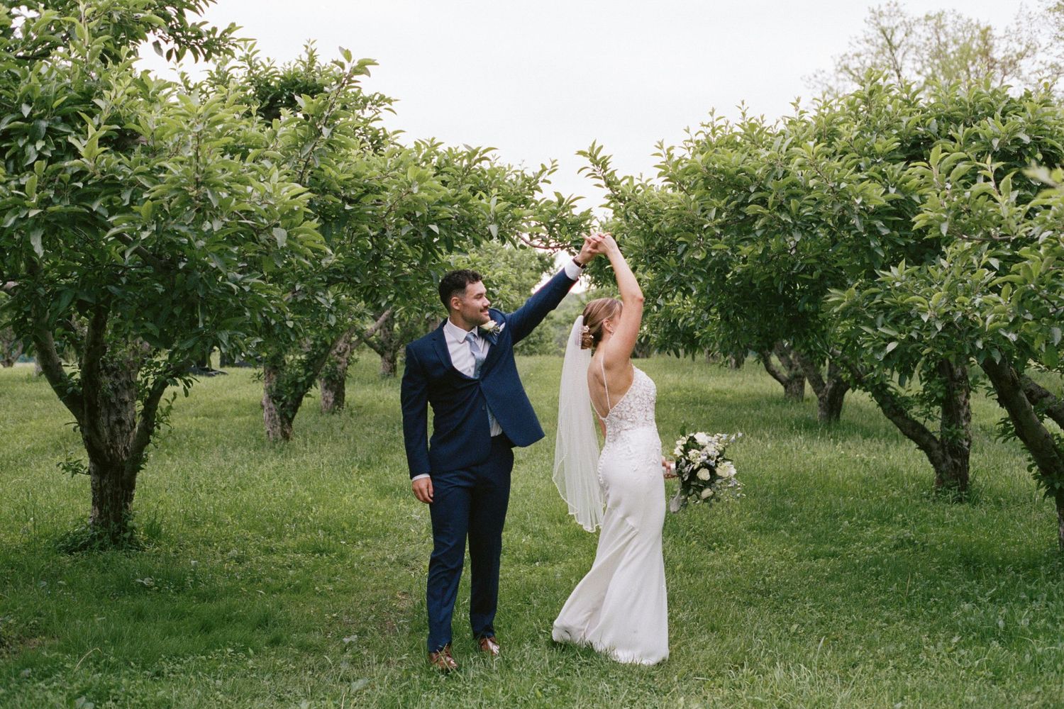 Bride and groom celebrate joyfully in lush orchard, groom raising bouquet under green trees.