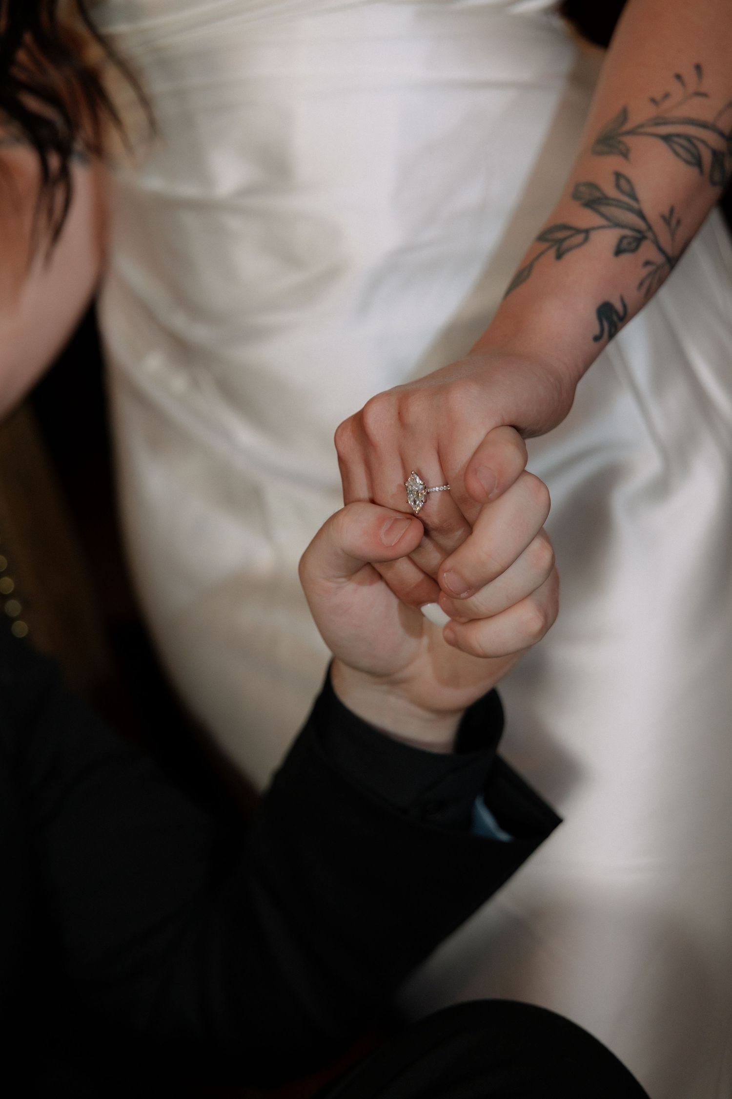 Close-up of bride and groom holding hands, showcasing a sparkling engagement ring and floral arm tattoo.