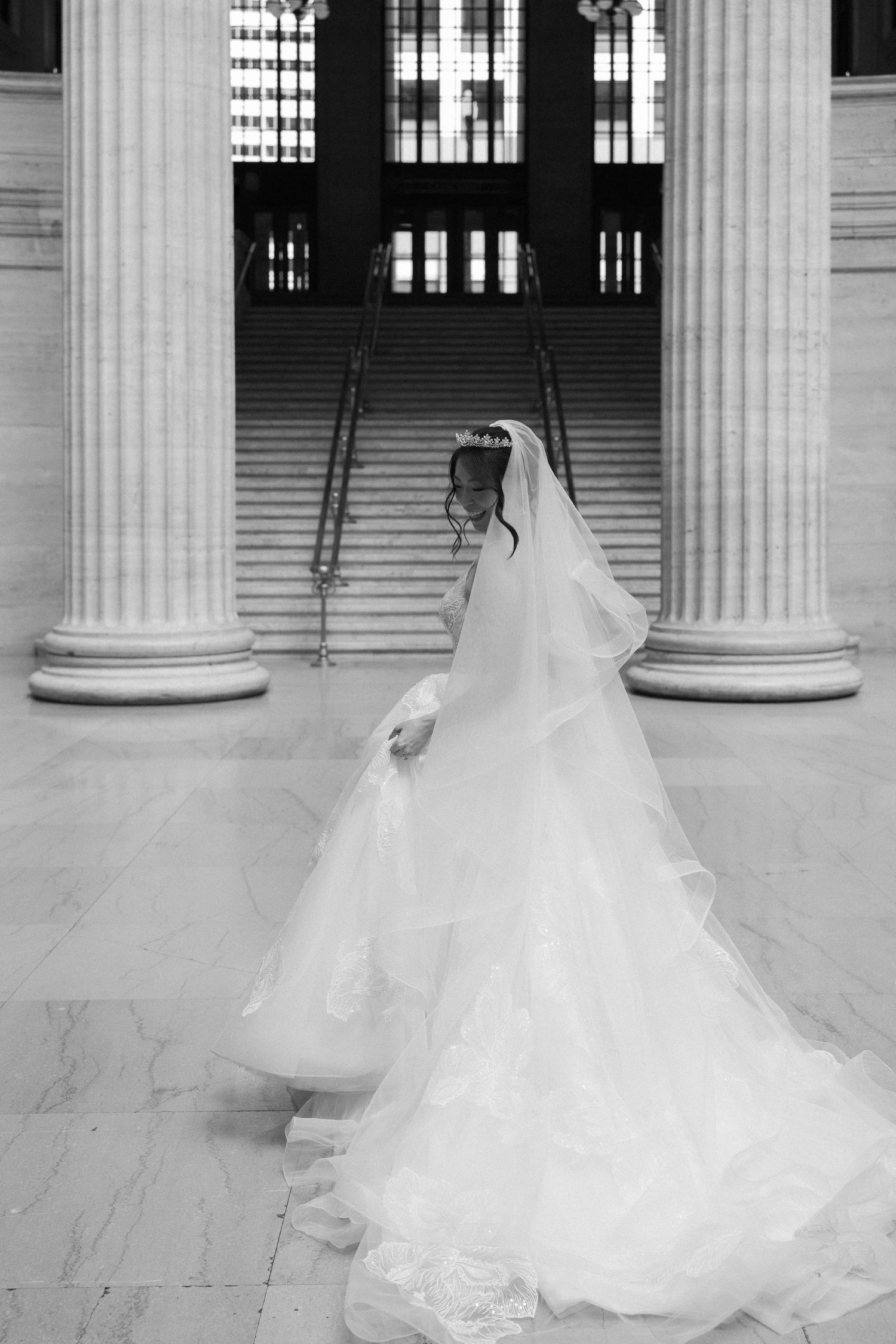 Black and white photo of bride in ballgown with long veil walking past marble columns.