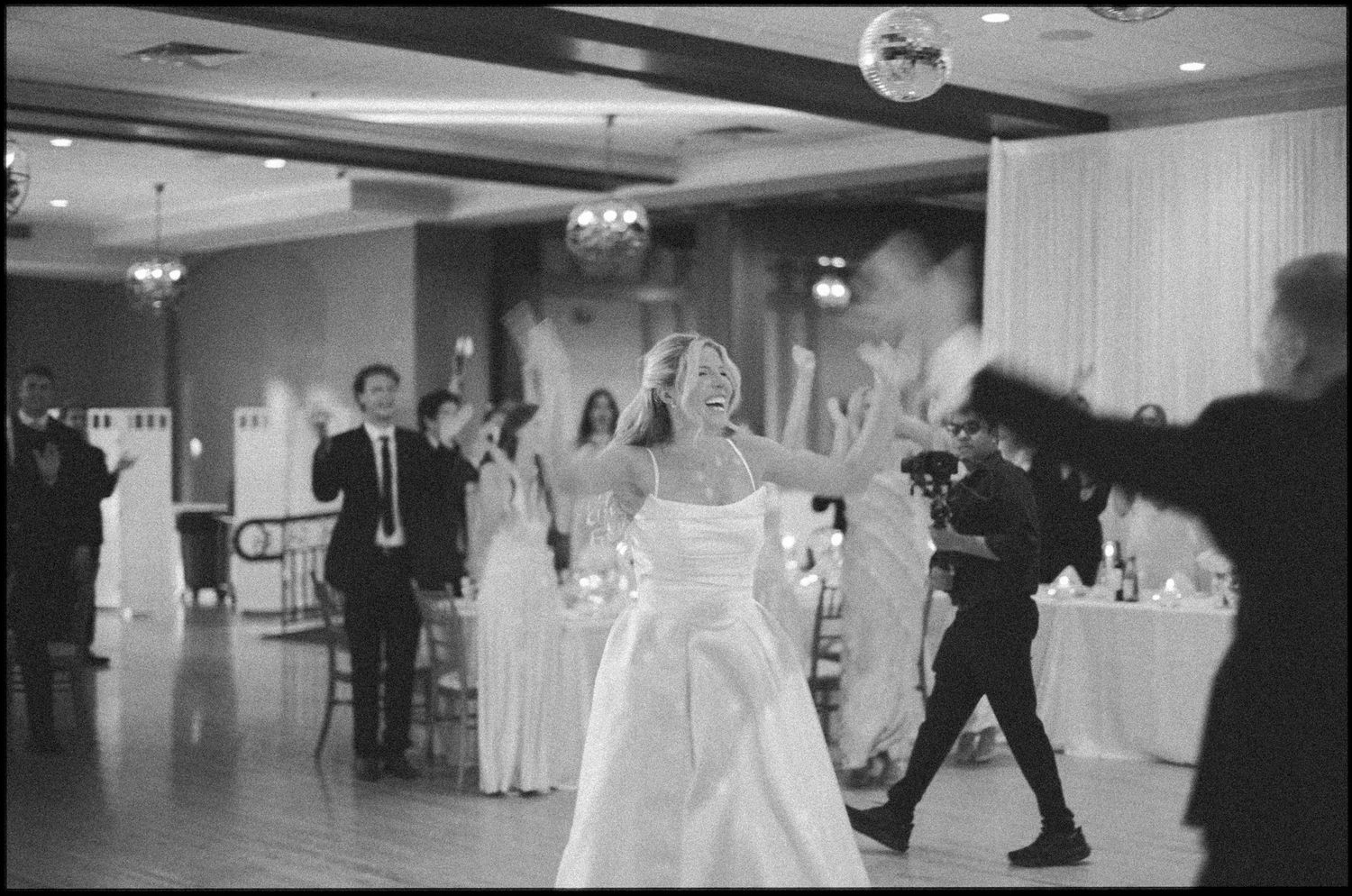 Bride laughing joyfully on wedding reception dance floor in white gown surrounded by guests in black and white photo.