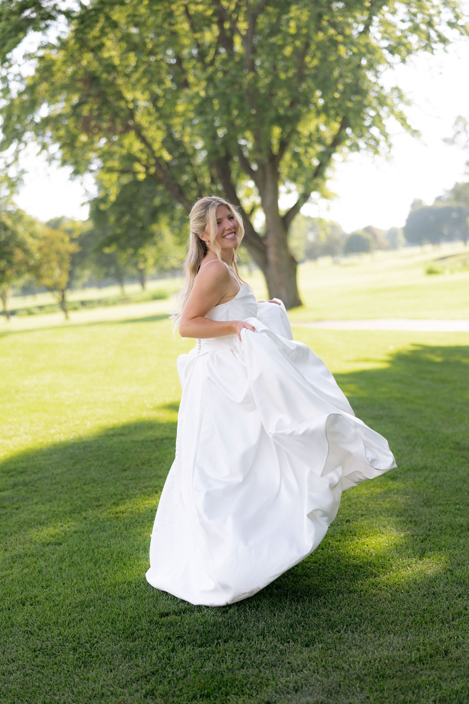 Joyful bride twirls in her white wedding gown on a sunlit green lawn surrounded by trees.