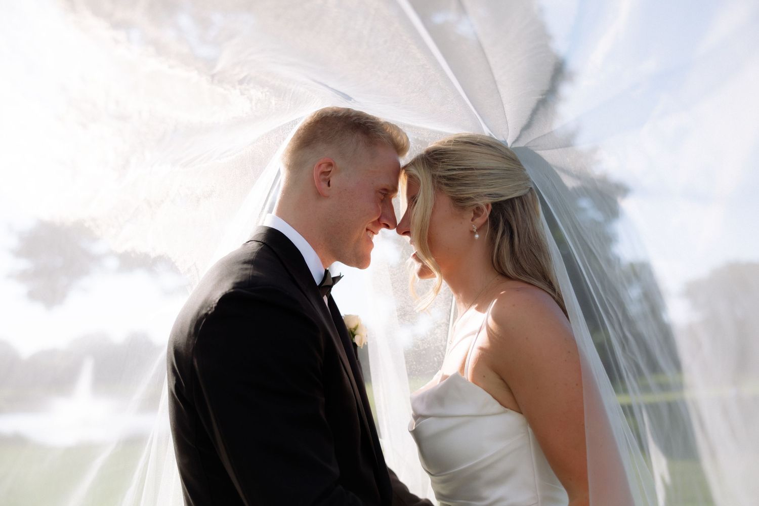 Bride and groom share romantic forehead touch under flowing veil in soft dreamy outdoor wedding portrait.