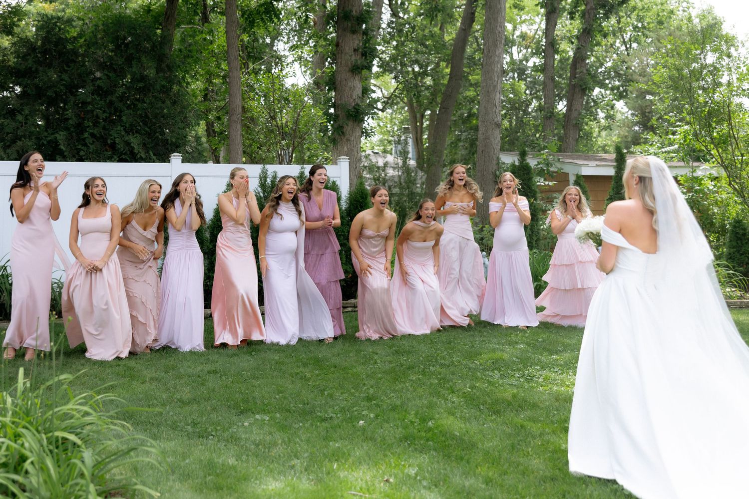Bride in white gown reveals herself to bridesmaids in pink dresses standing in a row outdoors.