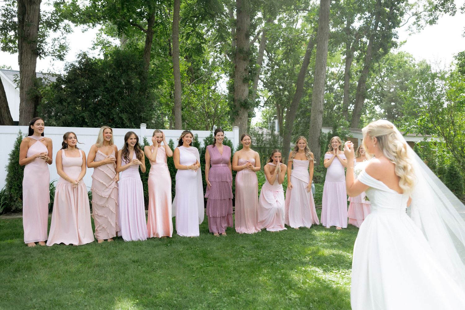 Bride reveals wedding dress to line of bridesmaids in pink gowns during outdoor first look.