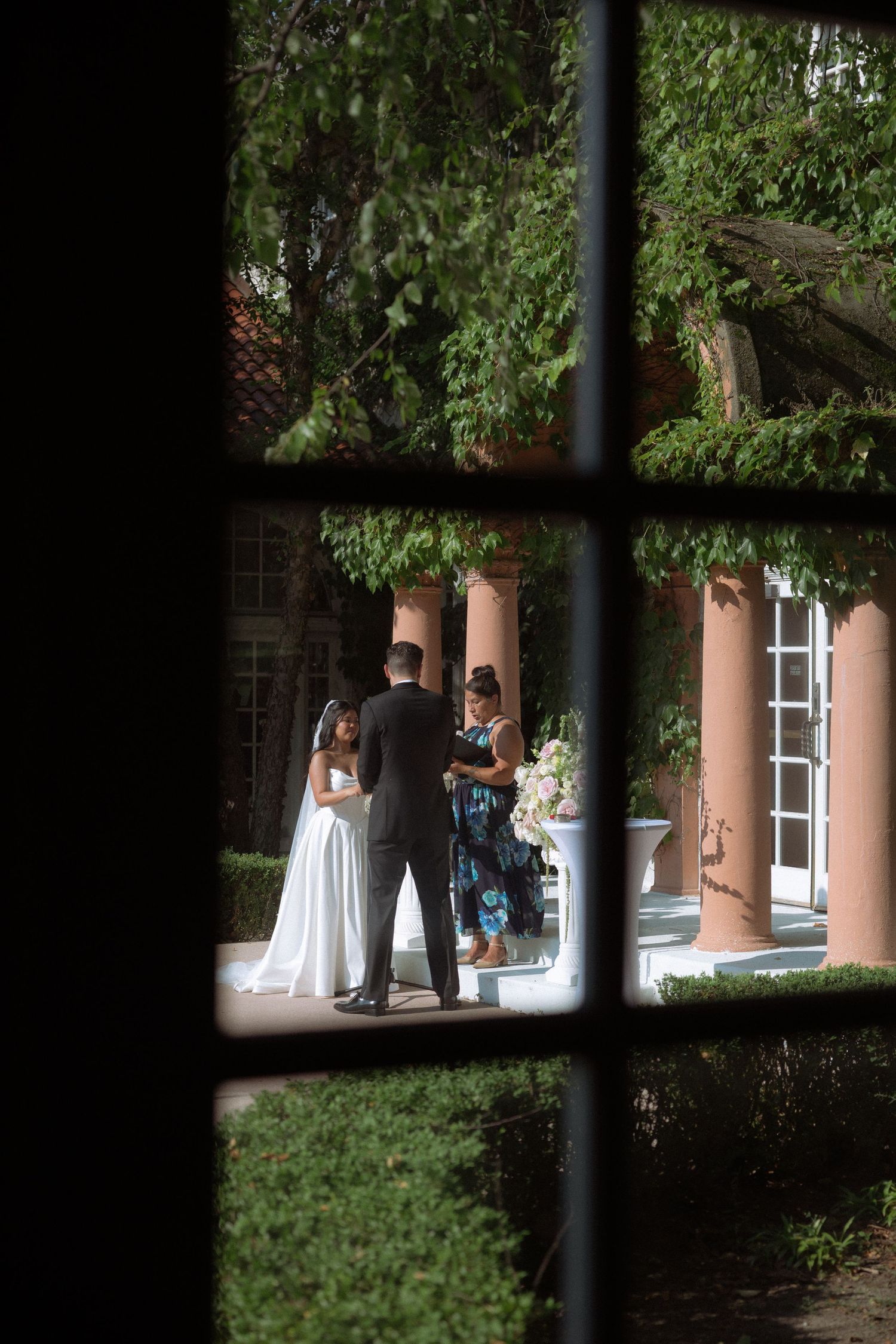 Bride and groom share intimate moment with officiant viewed through window panes in ivy-covered venue.