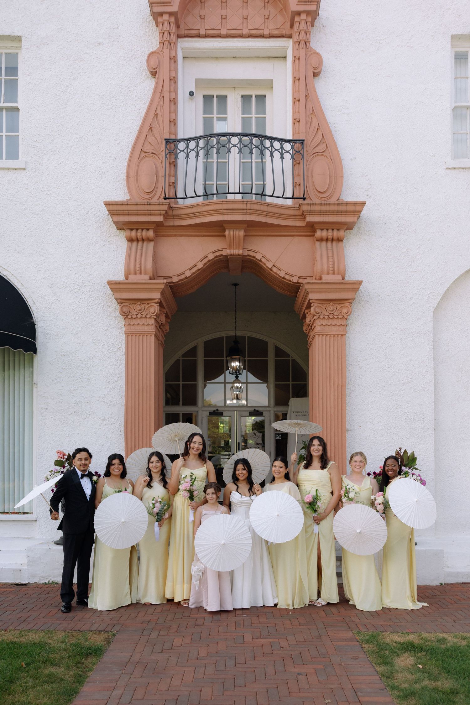 Bride with bridesmaids in yellow dresses holding white parasols posing in front of an elegant venue entrance.