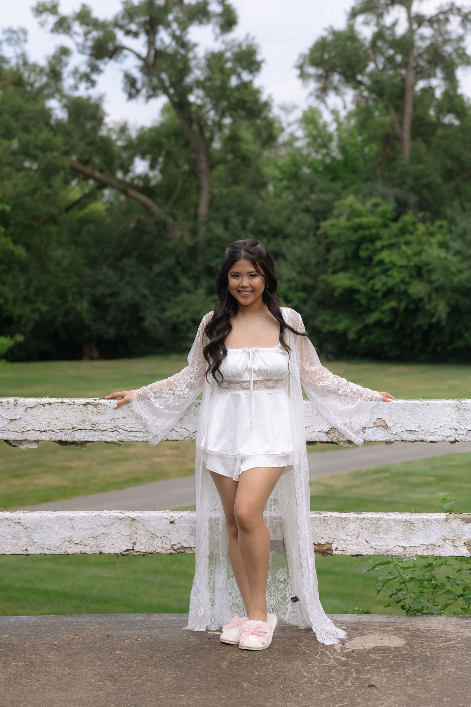 Young woman in white lace robe and romper posing by a white fence in a lush green park.