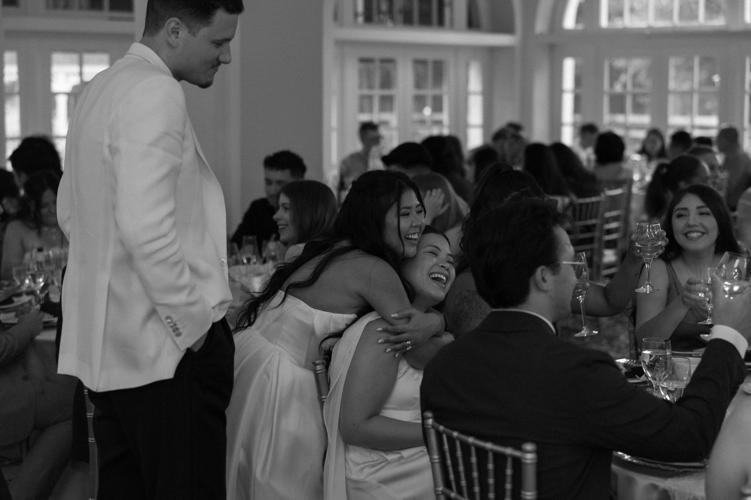 Black and white photo of bride hugging wedding guests during reception in elegant ballroom.