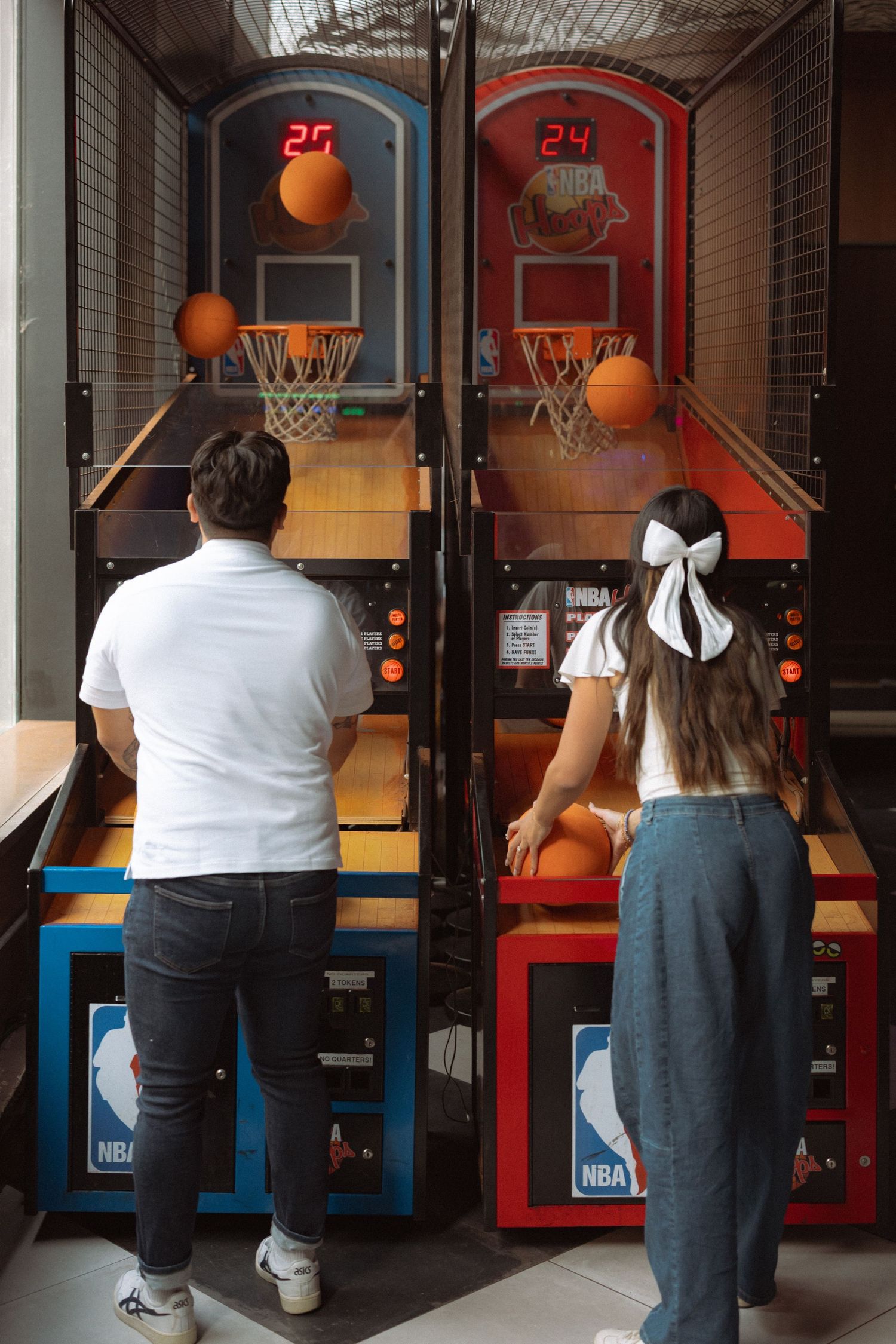 Two people playing NBA arcade basketball games side by side in an entertainment venue.