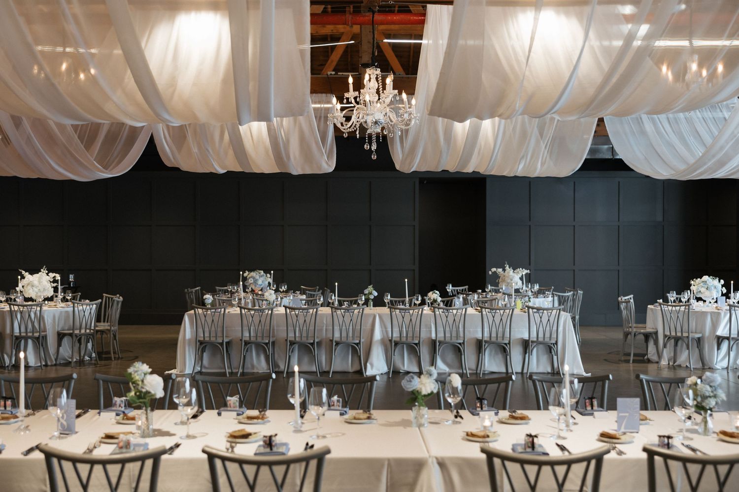 Elegant wedding reception hall with white draping, crystal chandelier, long tables with silver linens, and floral centerpieces.