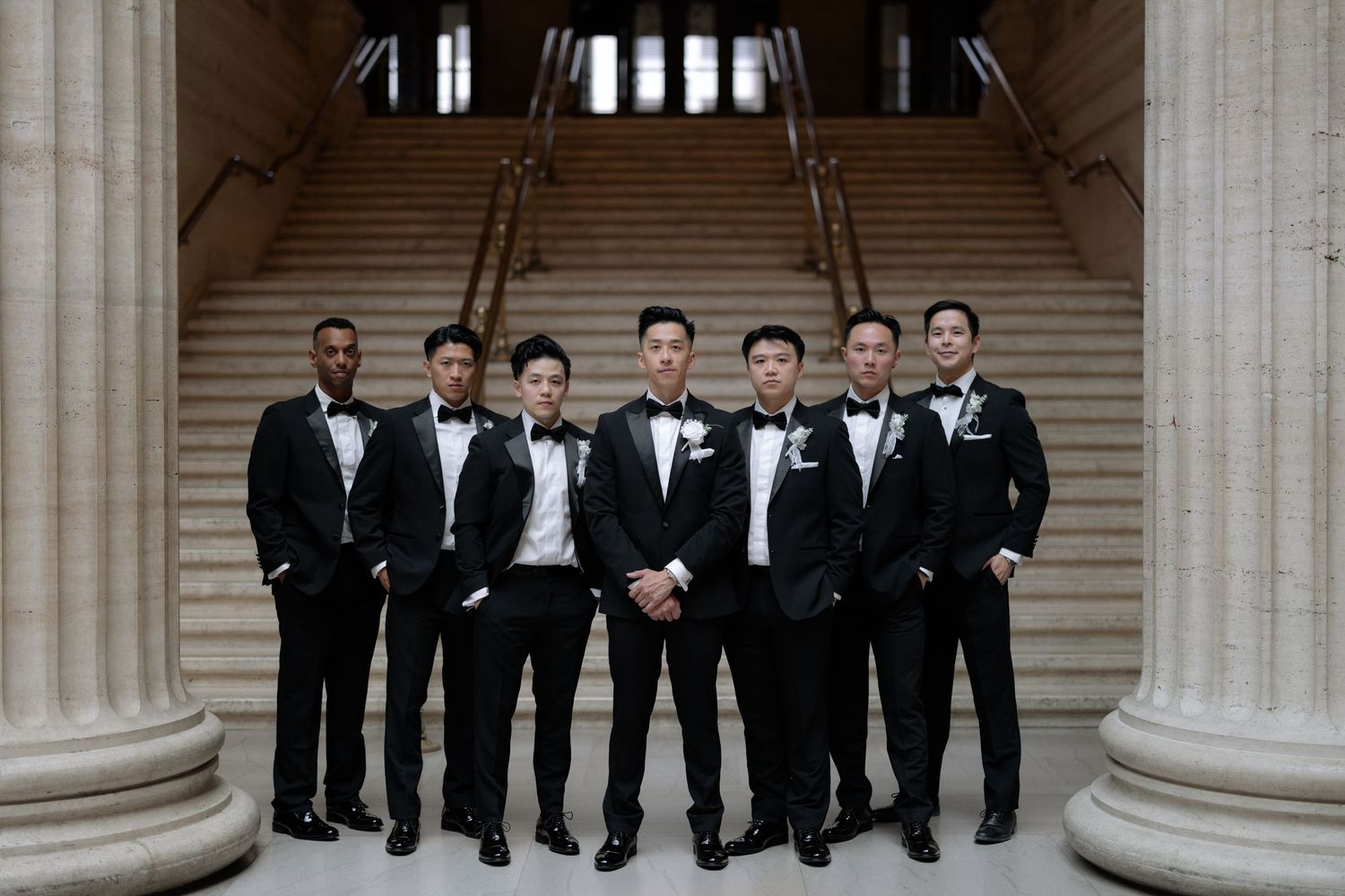 Groom and groomsmen pose in matching black tuxedos between stone columns on grand staircase.