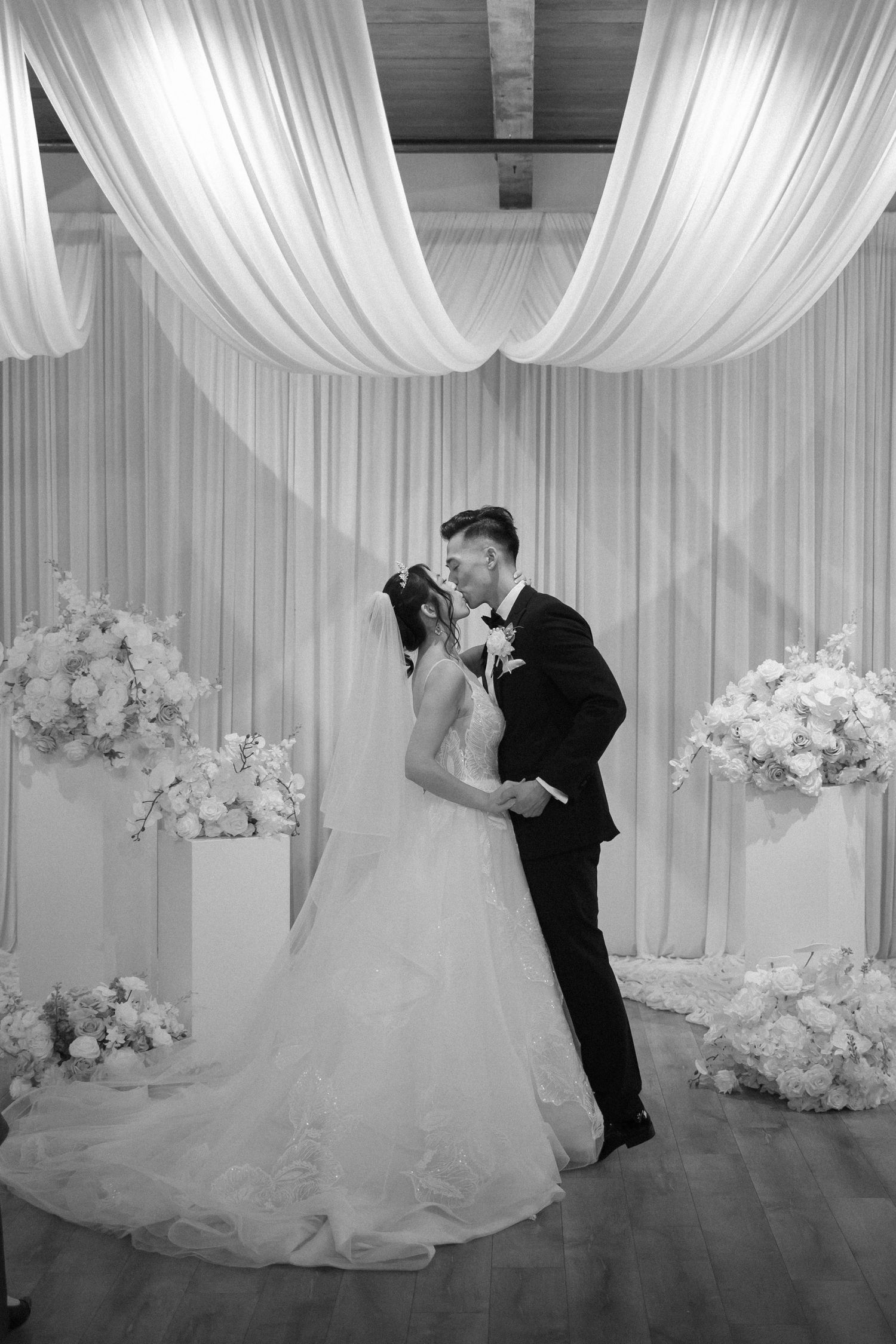 Bride and groom share first kiss at elegant wedding altar with white floral arrangements.