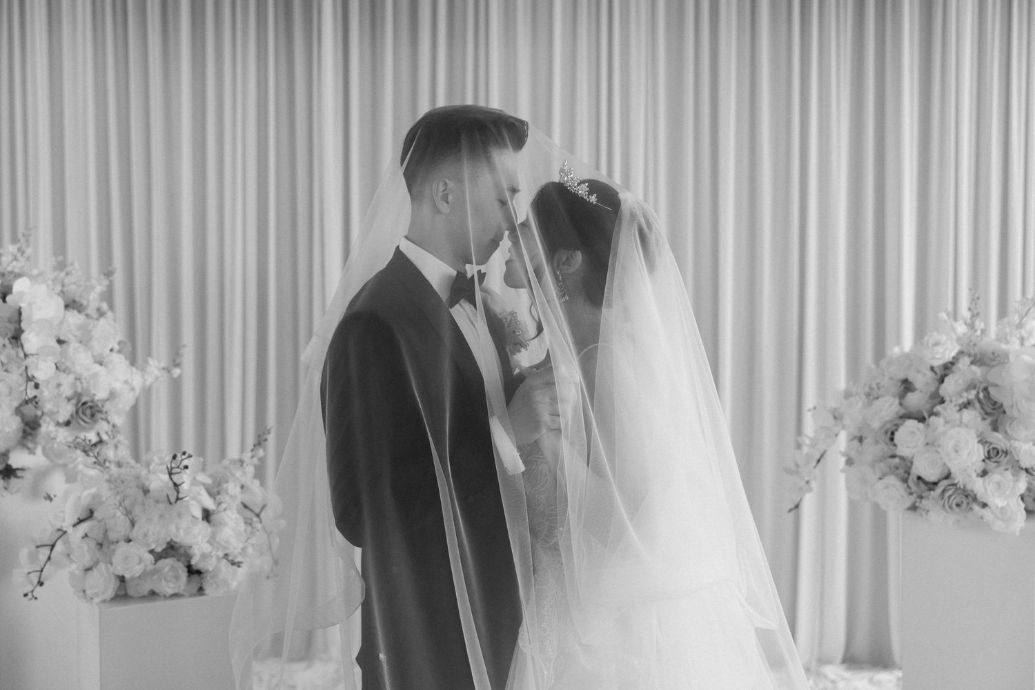 Black and white wedding photo of bride and groom sharing a kiss at the altar with floral arrangements.