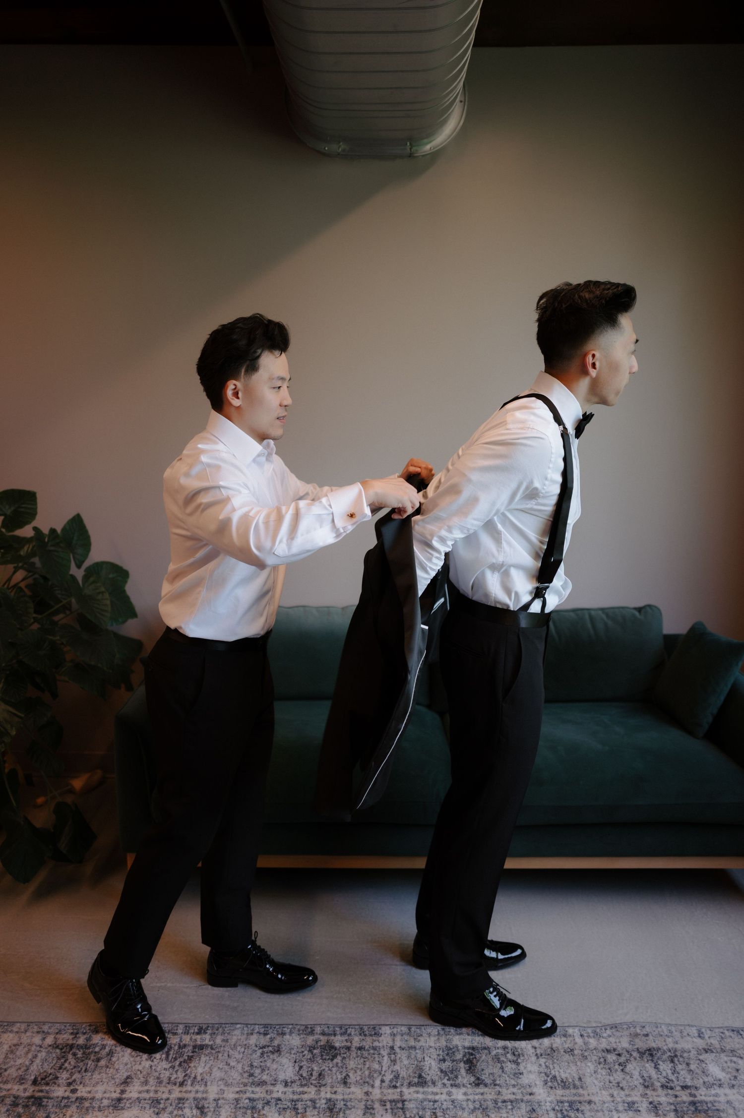A groomsman helps the groom put on his black suit jacket while both wear white dress shirts and black trousers.