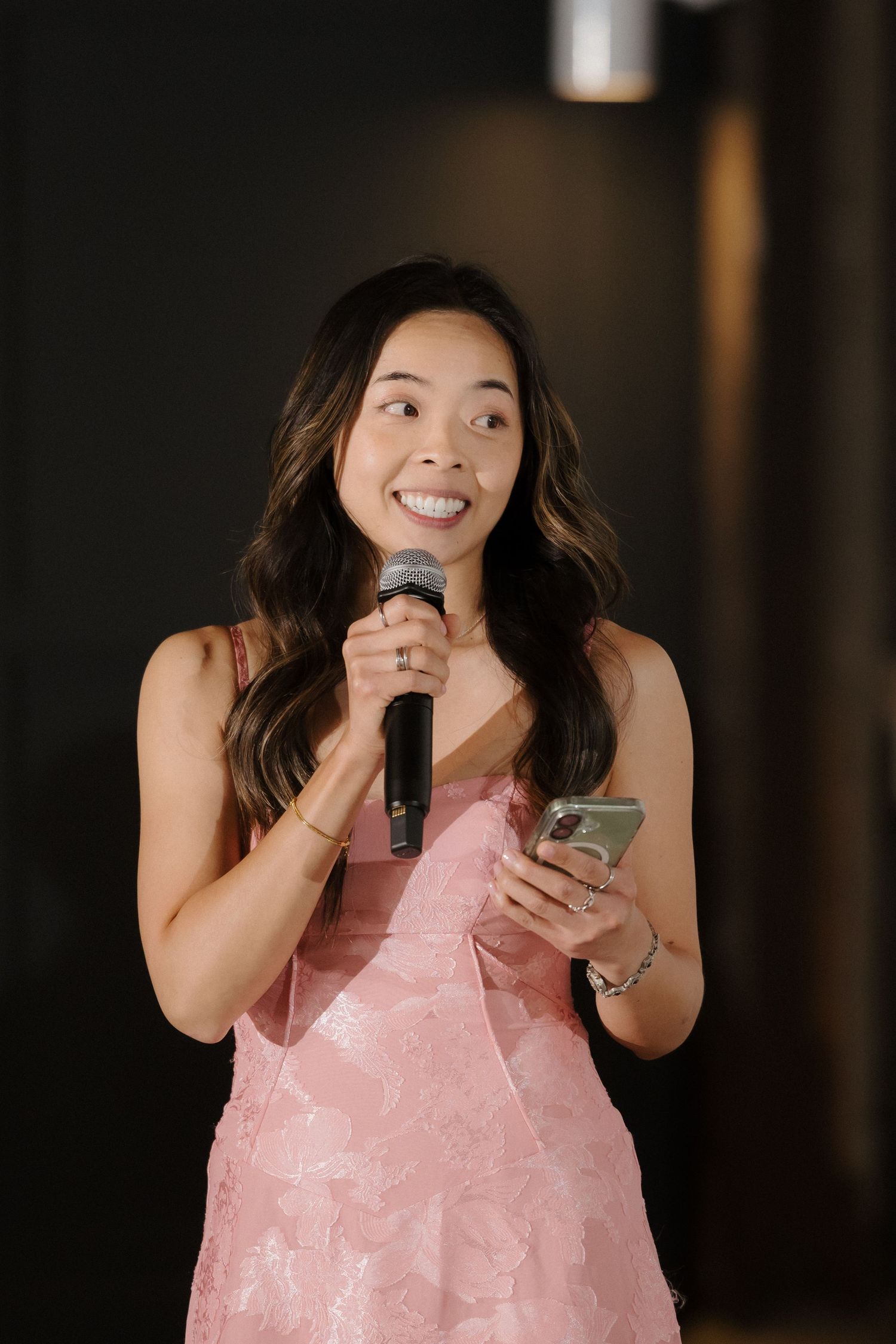 Woman in pink dress speaks into microphone holding phone at event.