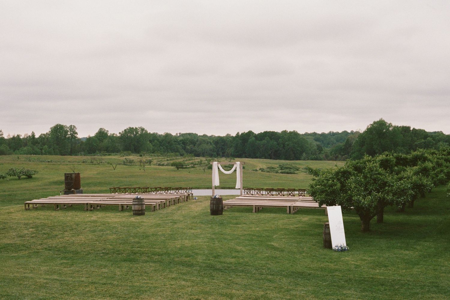 Outdoor wedding ceremony setup on wooden platform in lush green field with floral arch and rustic decor under overcast sky.