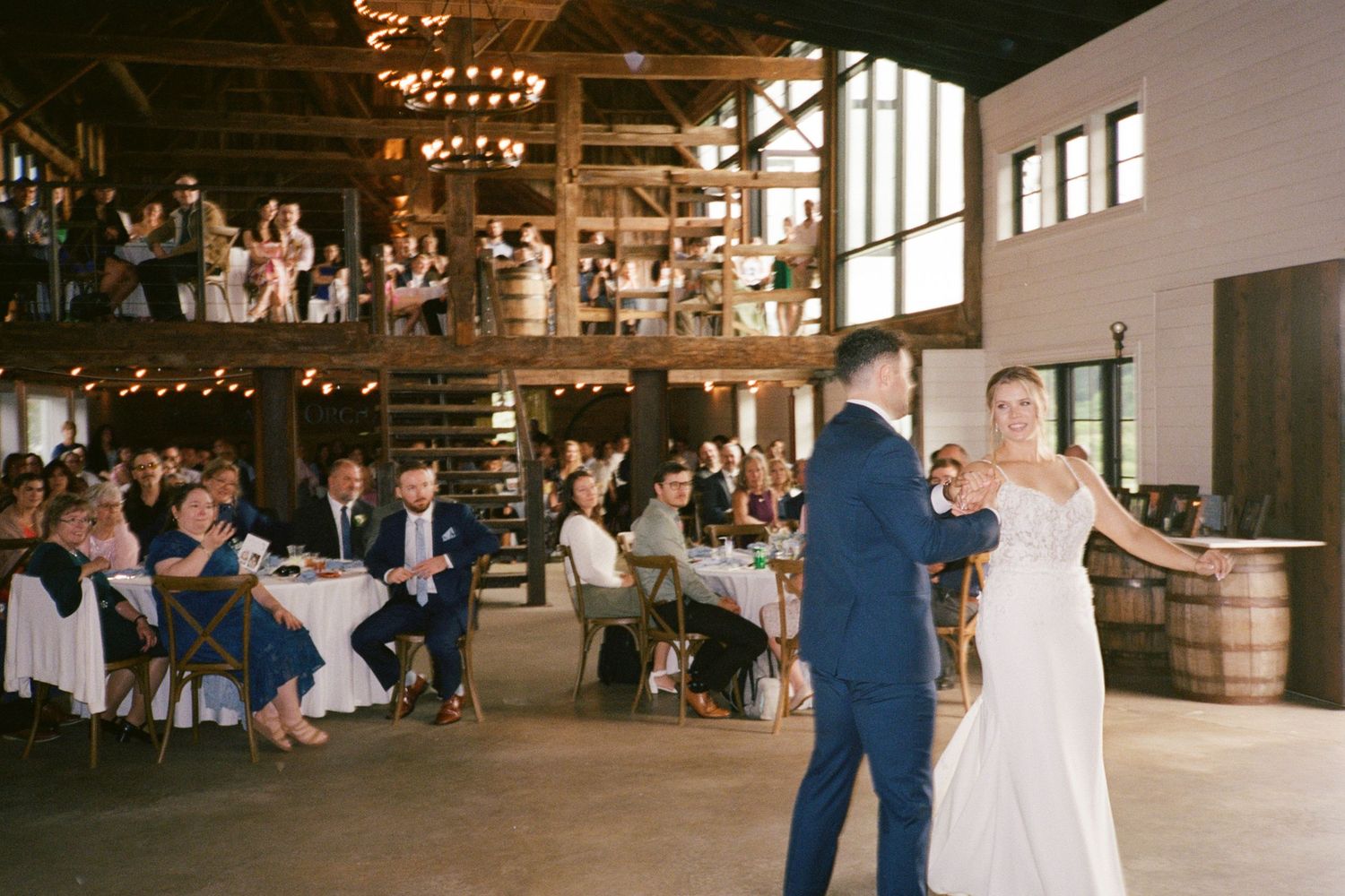 Bride and groom share first dance in rustic barn wedding venue with guests seated at round tables on upper level.