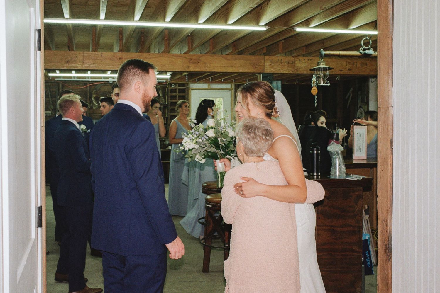 Bride holding bouquet walks with bridesmaid in blush dress toward groom in navy suit inside rustic barn wedding venue.