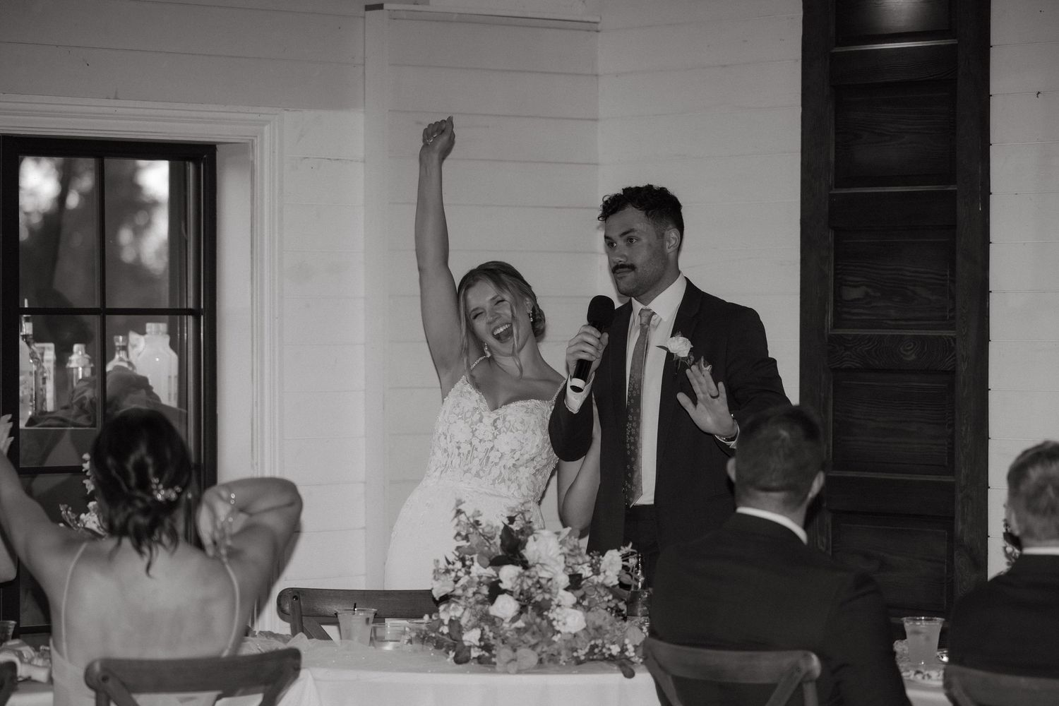 Black and white photo of bride and groom celebrating at wedding reception with microphone.