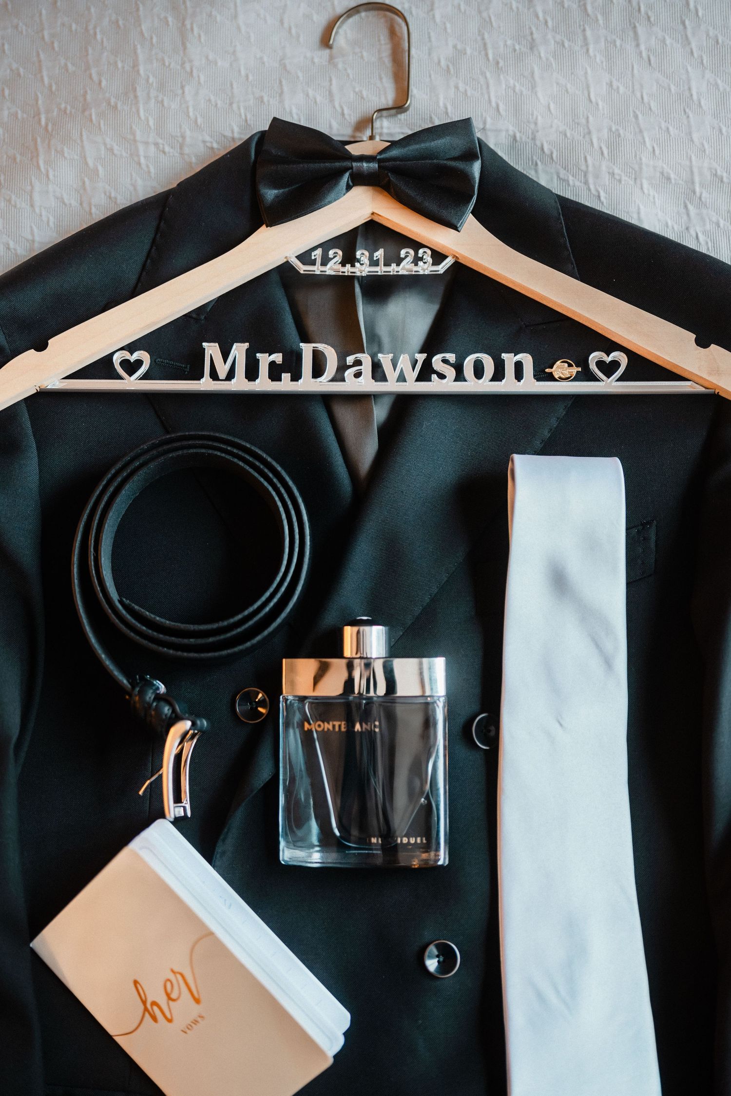 Groom's wedding flat lay featuring a black tuxedo, bow tie, belt, cologne, and Mr. Dawson personalized hanger.