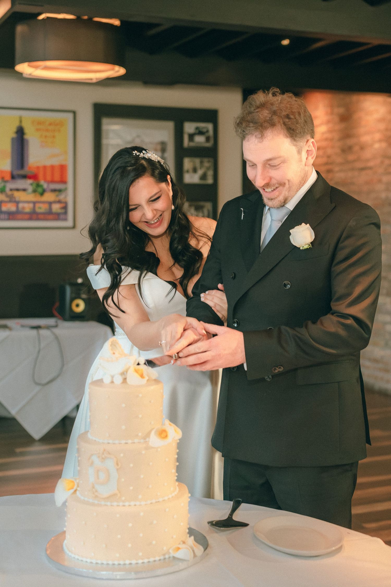 Smiling bride and groom cut elegant three-tier white wedding cake at reception.