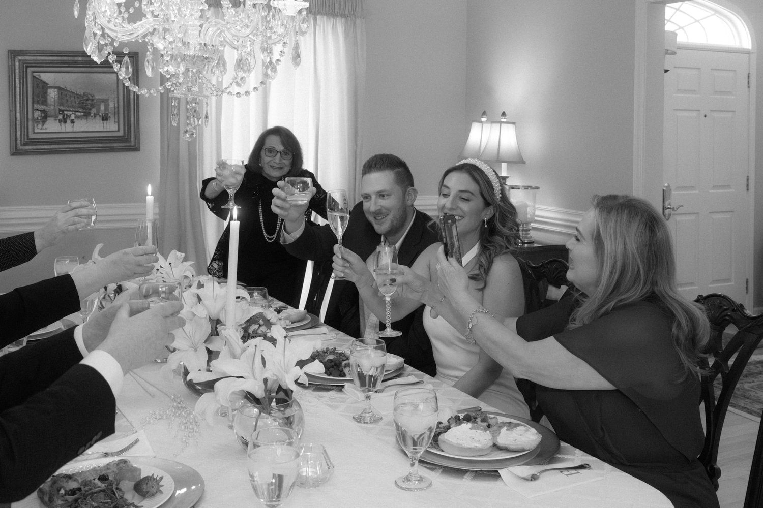 Black and white photo of wedding party raising glasses in a joyful toast at the reception dinner table.