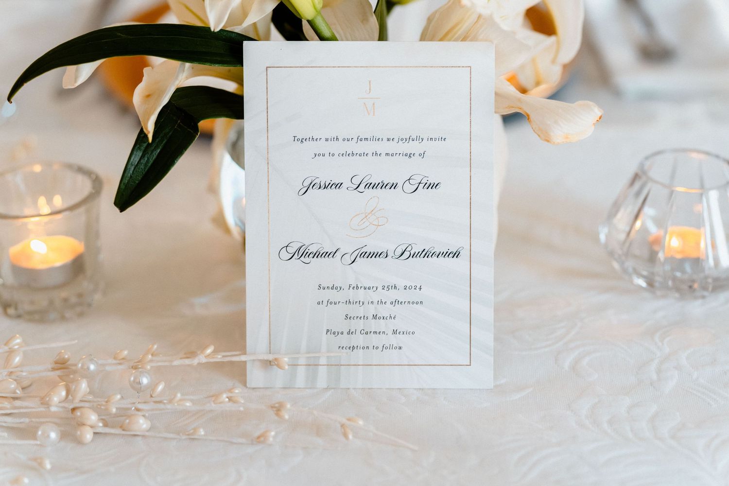 Elegant wedding invitation with gold border displayed on a white tablecloth with candles and white lilies.