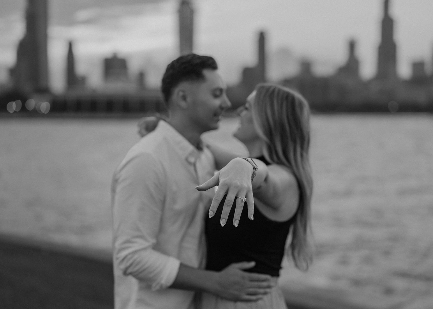 Black and white photo of couple embracing near water, woman showing off engagement ring.