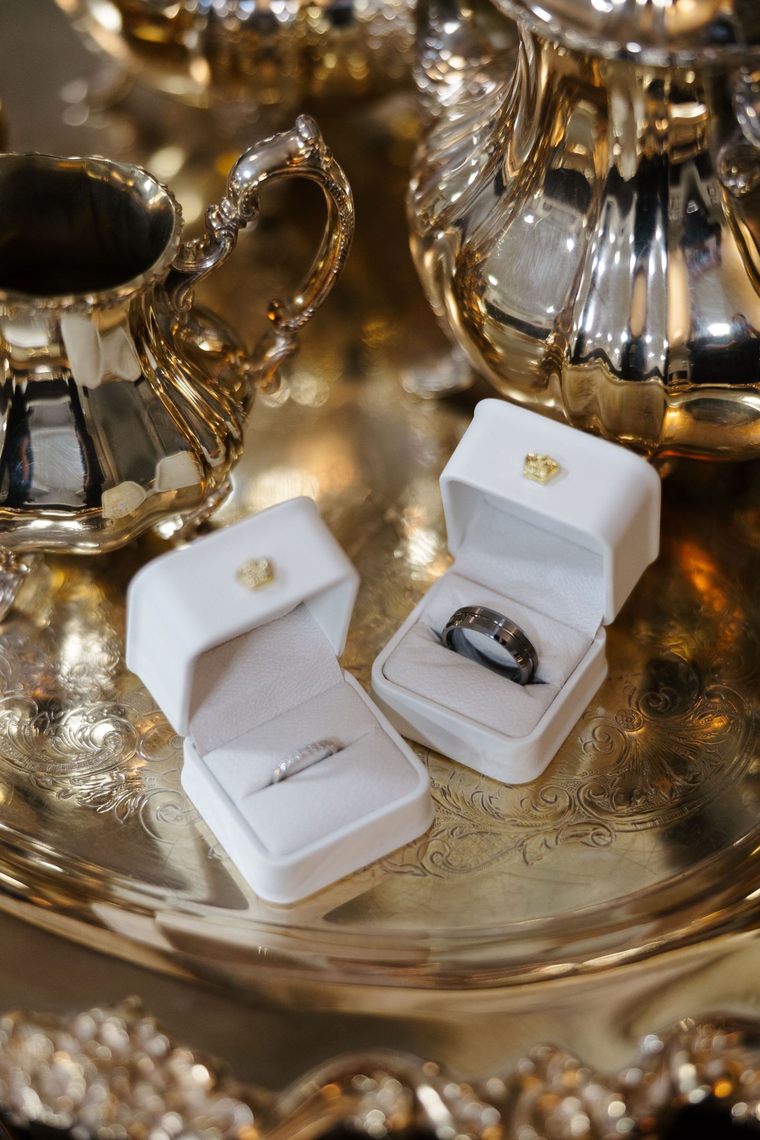 Two white velvet ring boxes with wedding bands displayed on an ornate silver tray with elegant tea service.