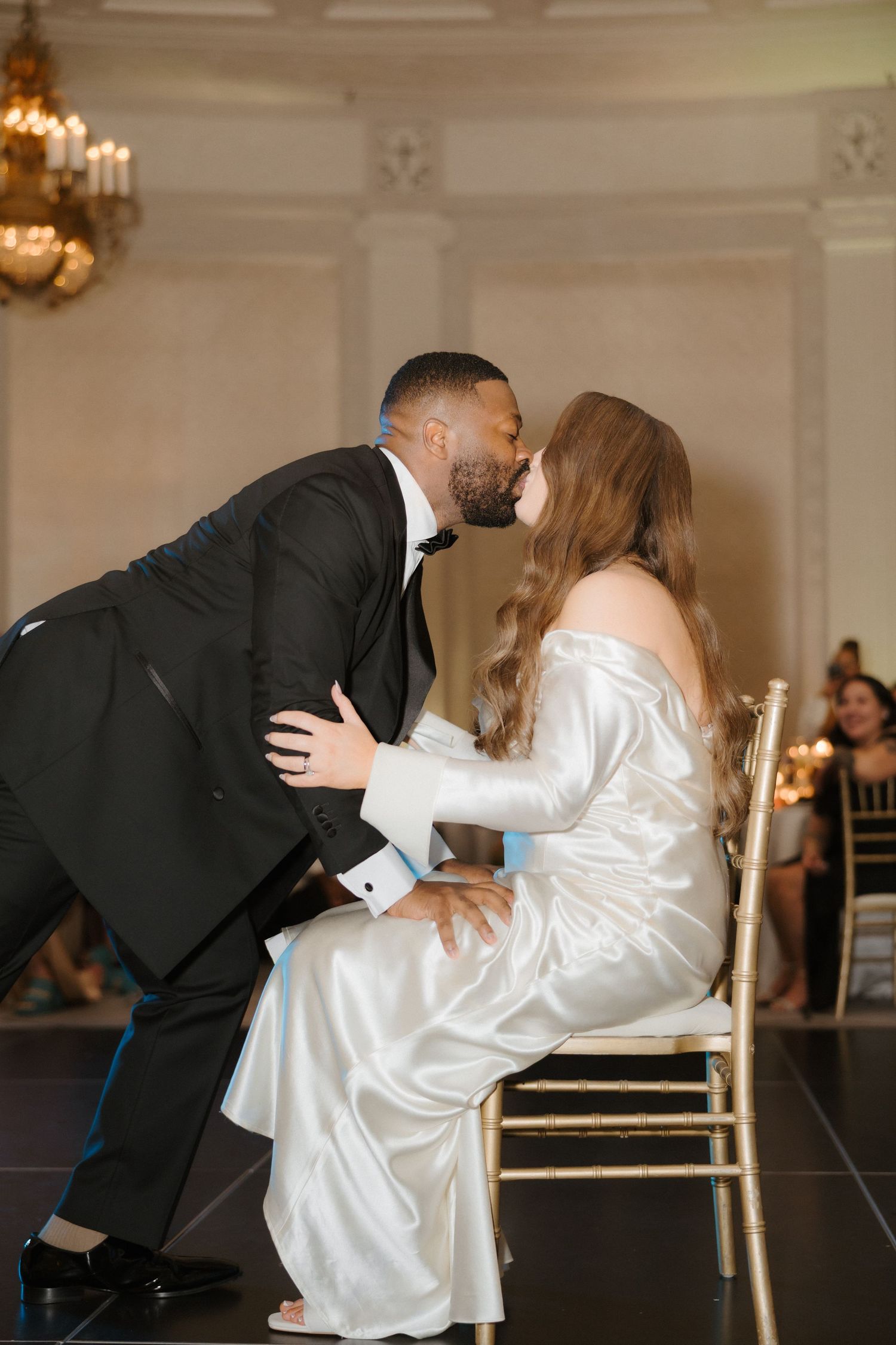 Bride in white satin gown shares a kiss with groom in black tuxedo during wedding reception chair dance.