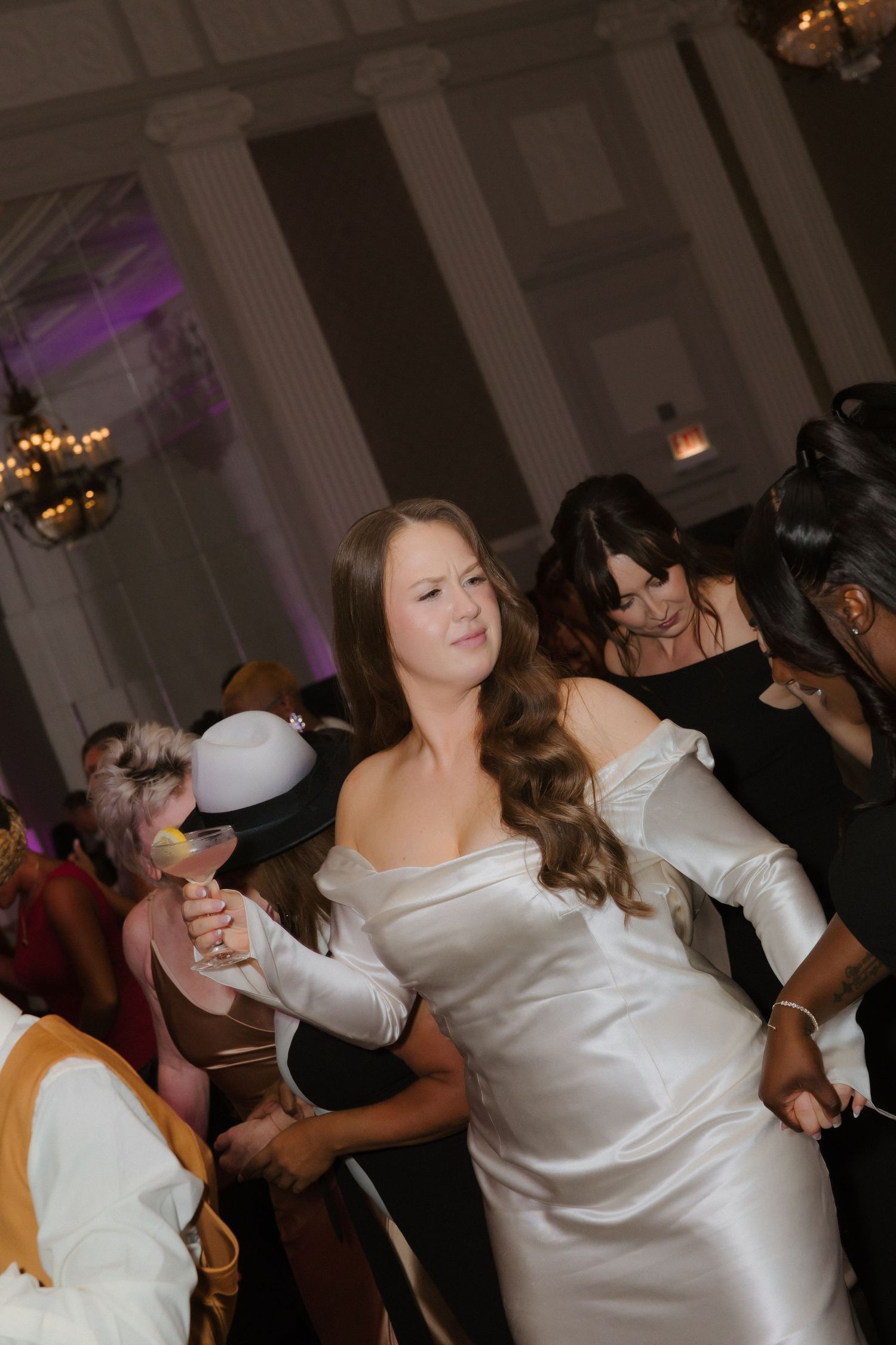Bride in white off-shoulder gown dancing joyfully with guests at elegant wedding reception venue.