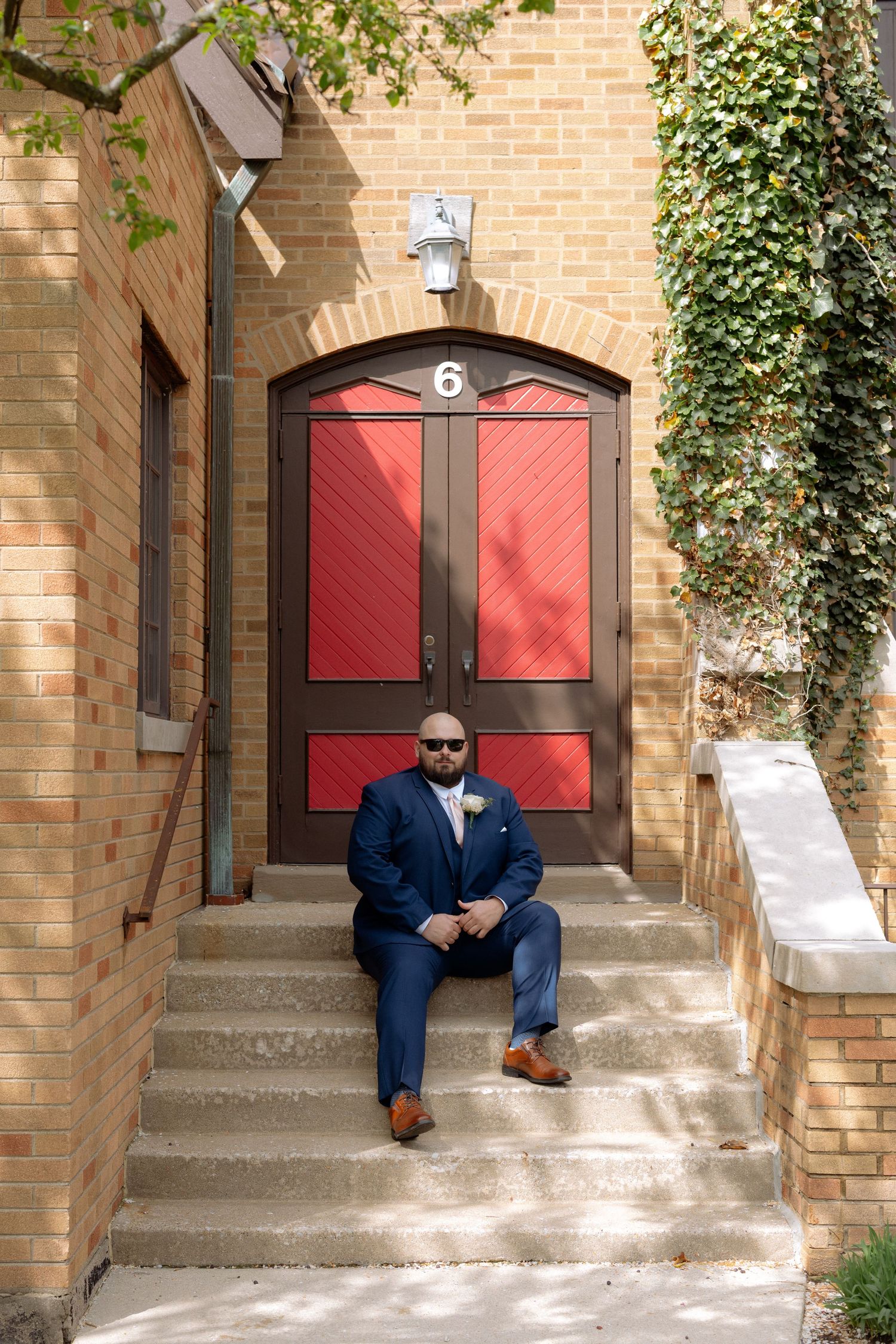 Groom in navy suit sitting on steps before a red arched door numbered 6, surrounded by ivy-covered brick walls.