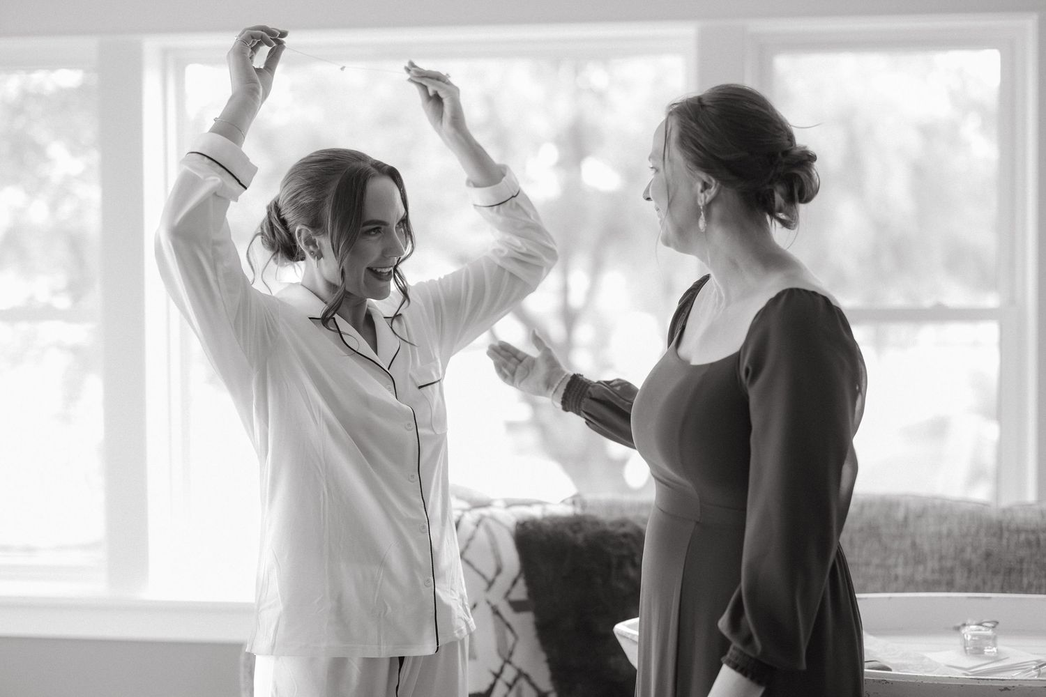 Black and white photo of bride in white pajamas dancing with arms raised while bridesmaid watches indoors.