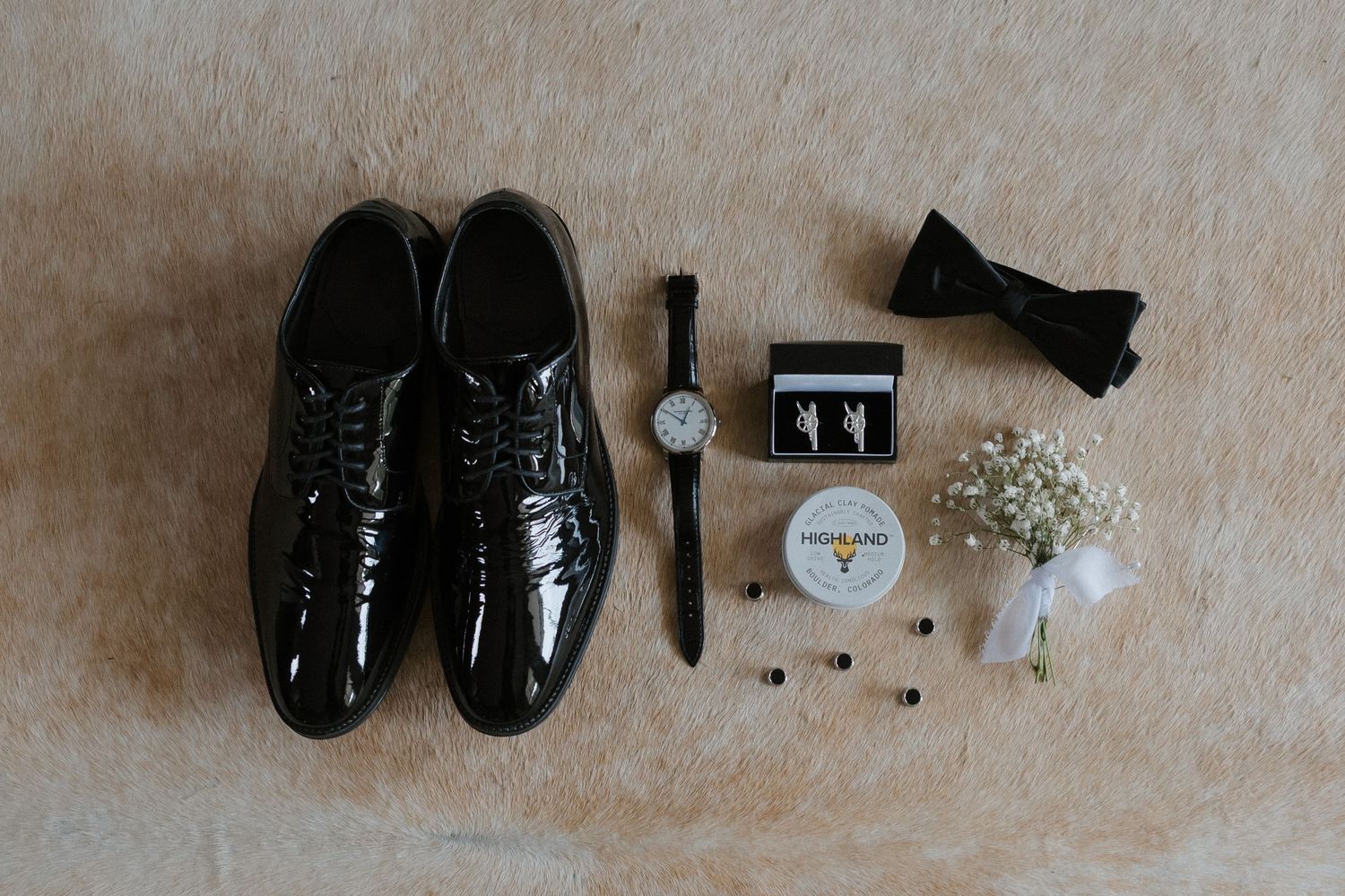 Flat lay of groom's wedding accessories including black patent shoes, watch, bow tie, and boutonniere.