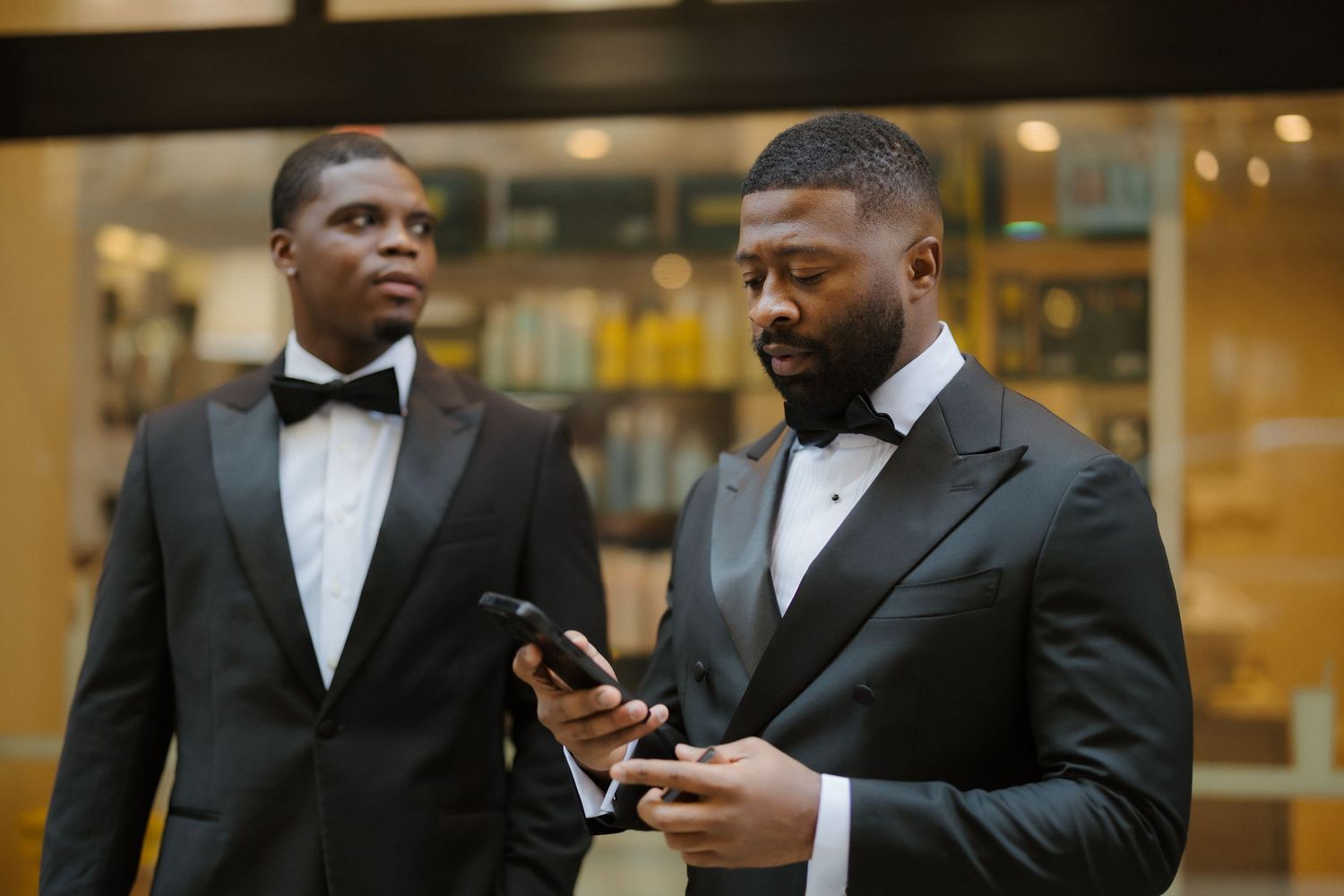 Two men in black tuxedos with bow ties, one checking a smartphone, standing near a glass storefront.