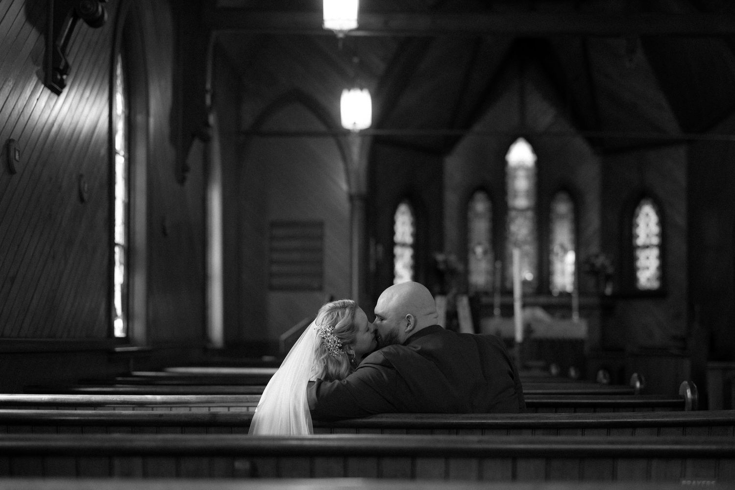 Black and white photo of a bride and groom sharing an intimate moment in a church pew before a stained glass altar.
