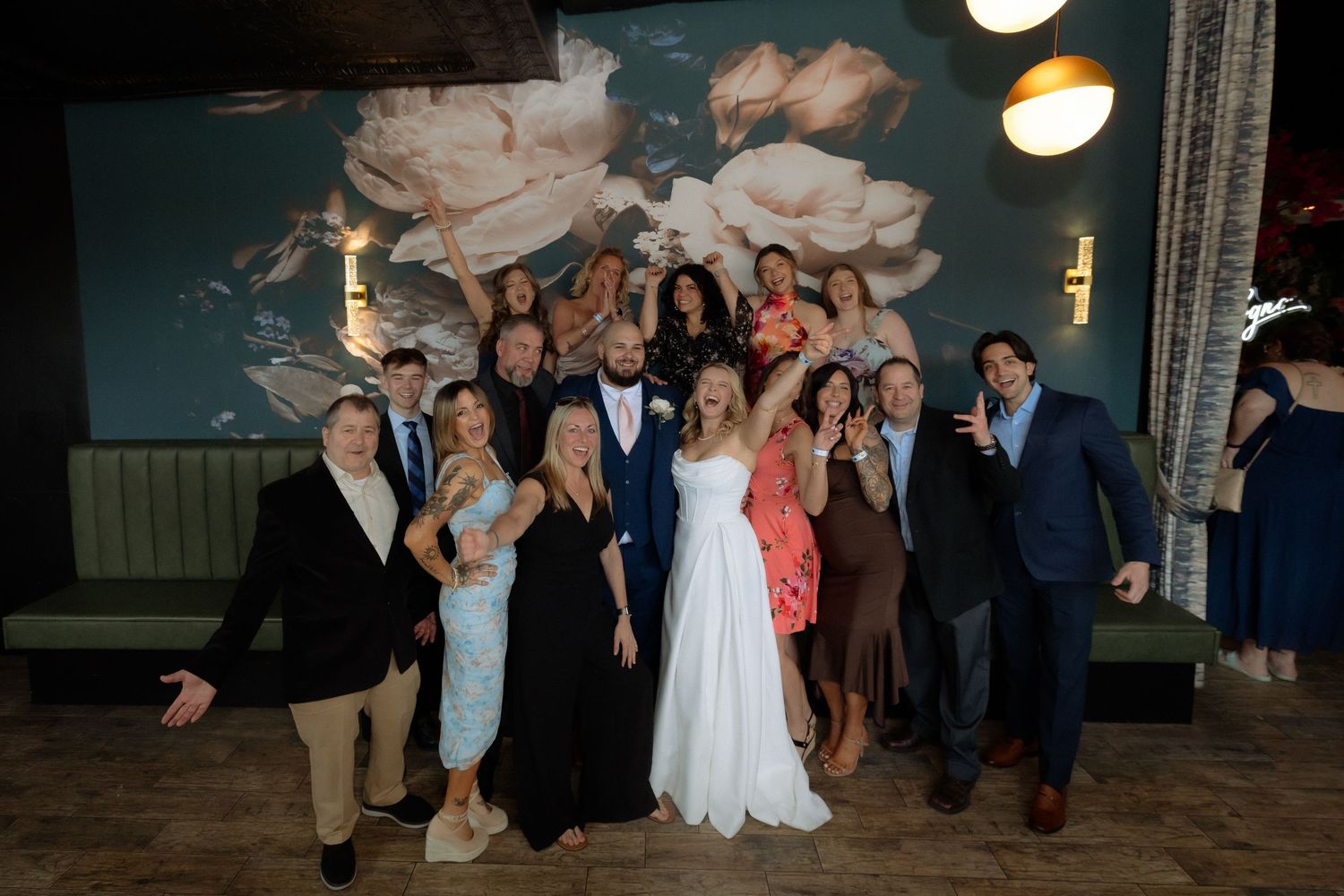 Wedding couple poses with joyful group of guests in front of floral mural at elegant reception venue.
