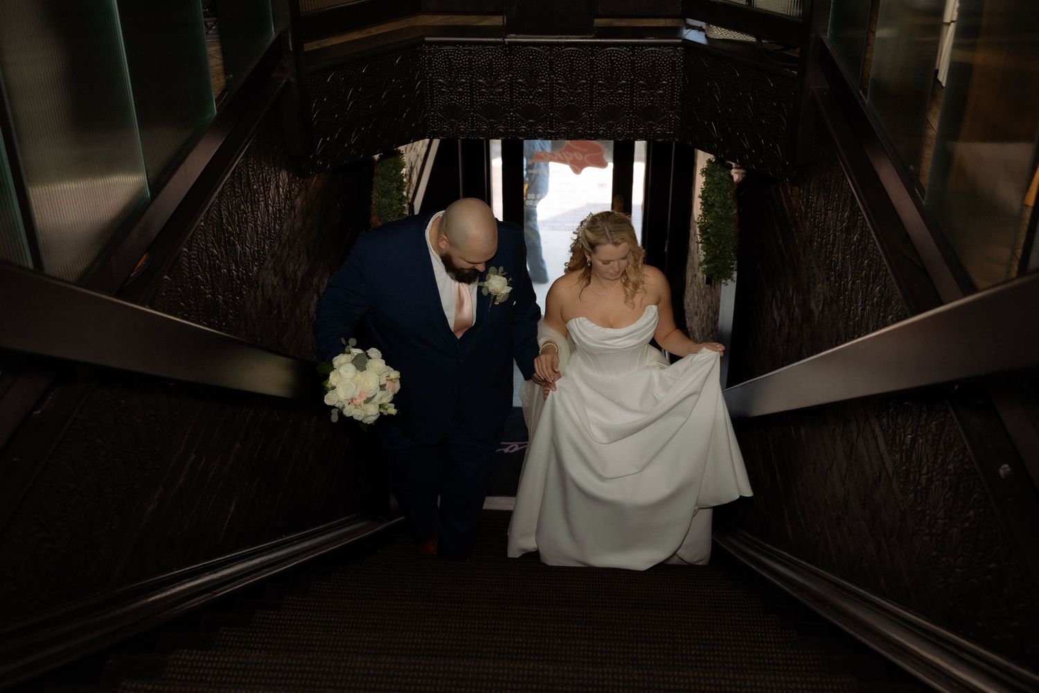 Bride and groom descend elegant dark wood staircase, bride in white gown holding bouquet of white flowers at wedding venue.