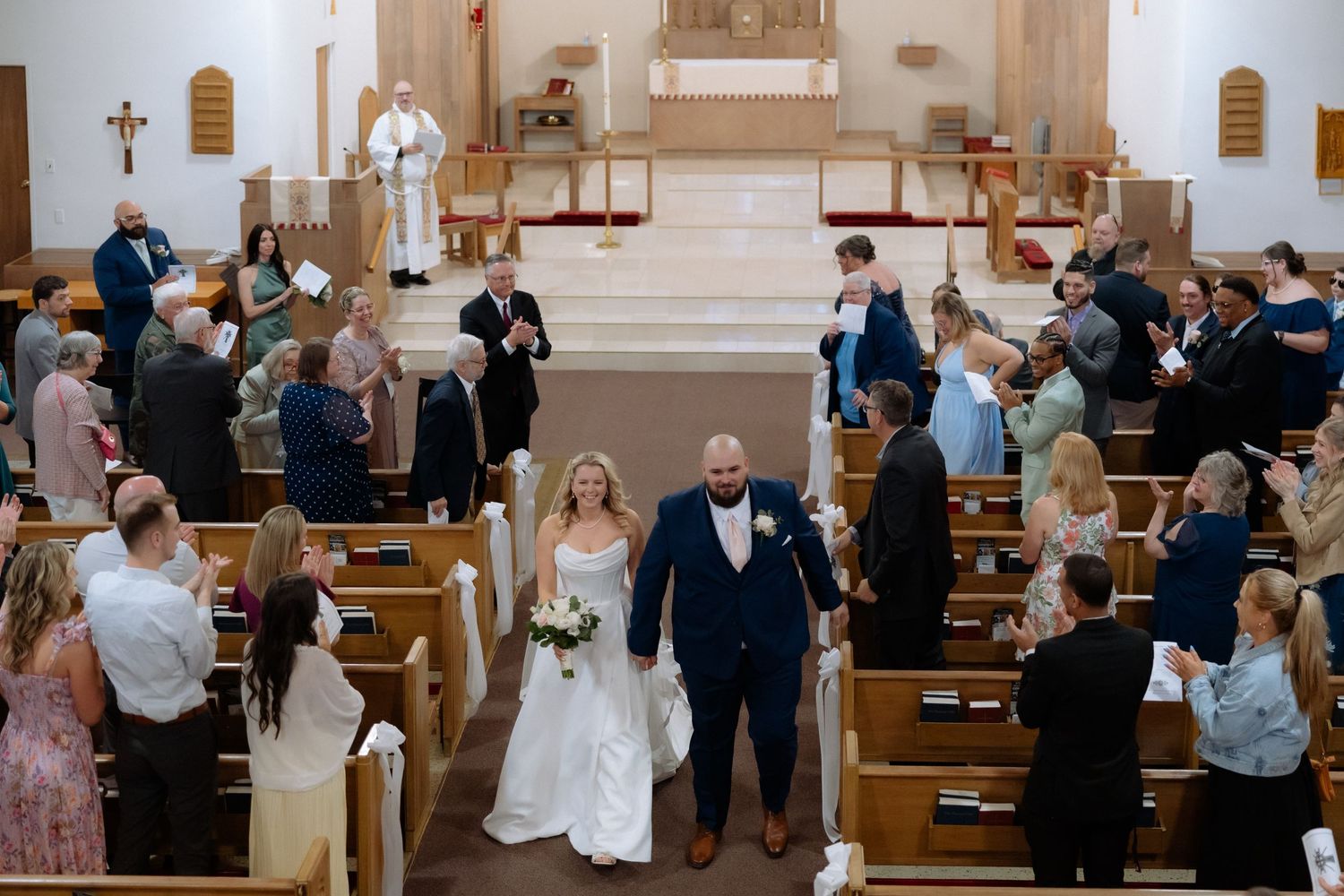 Bride and groom walk down church aisle after wedding ceremony, guests standing in wooden pews, priest at altar.